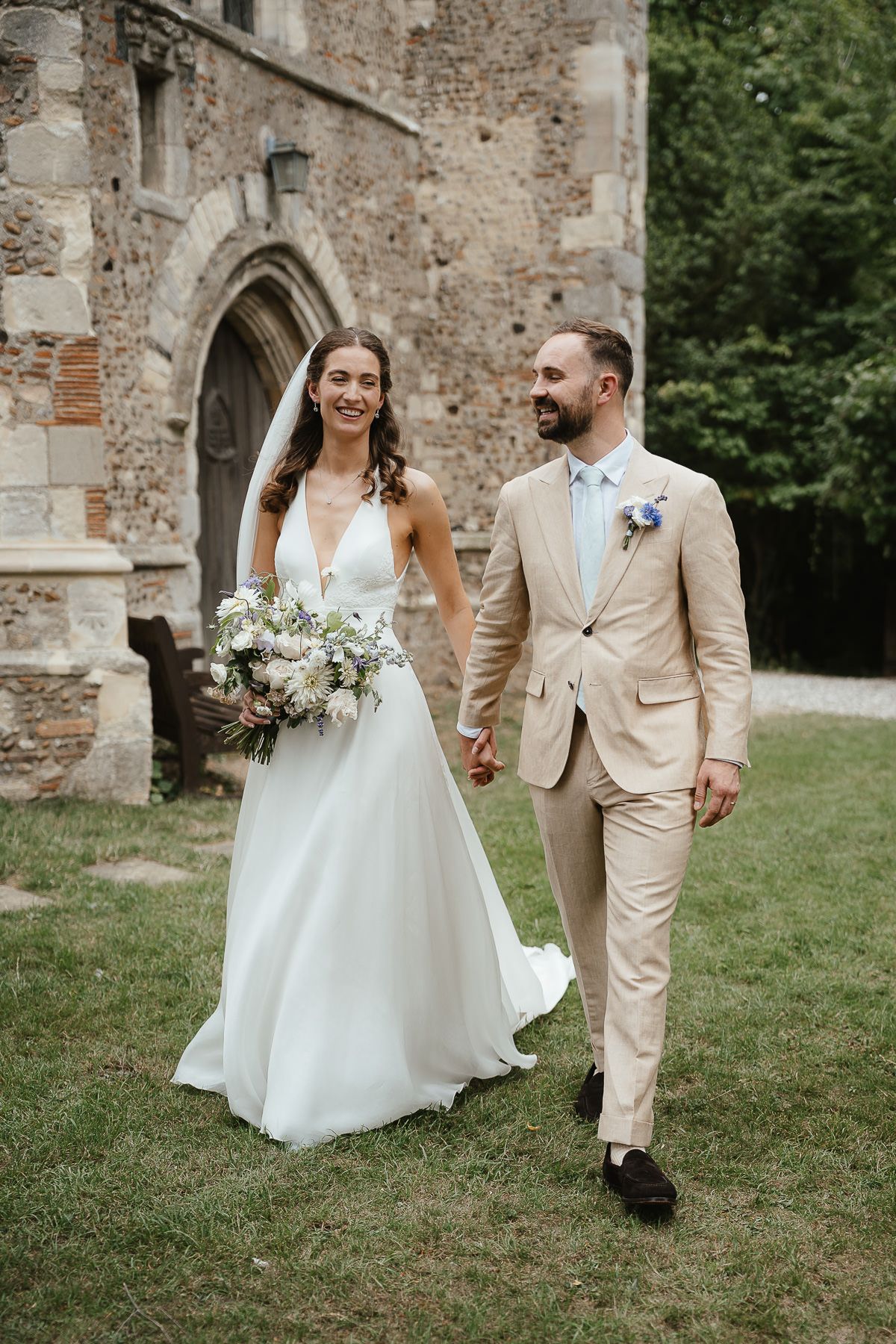 Newlywed couple walking joyfully together outside historic stone church ruins in casual summer wedding attire.