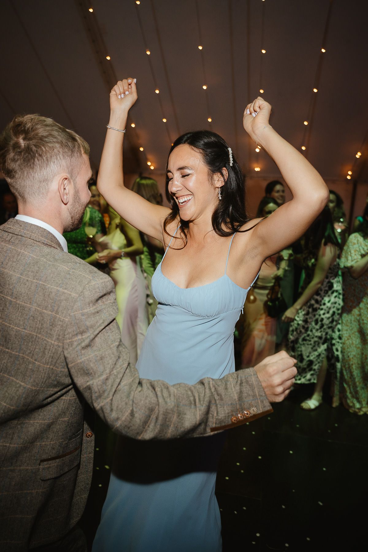 Two people dancing energetically at a wedding reception with string lights and guests in the background.