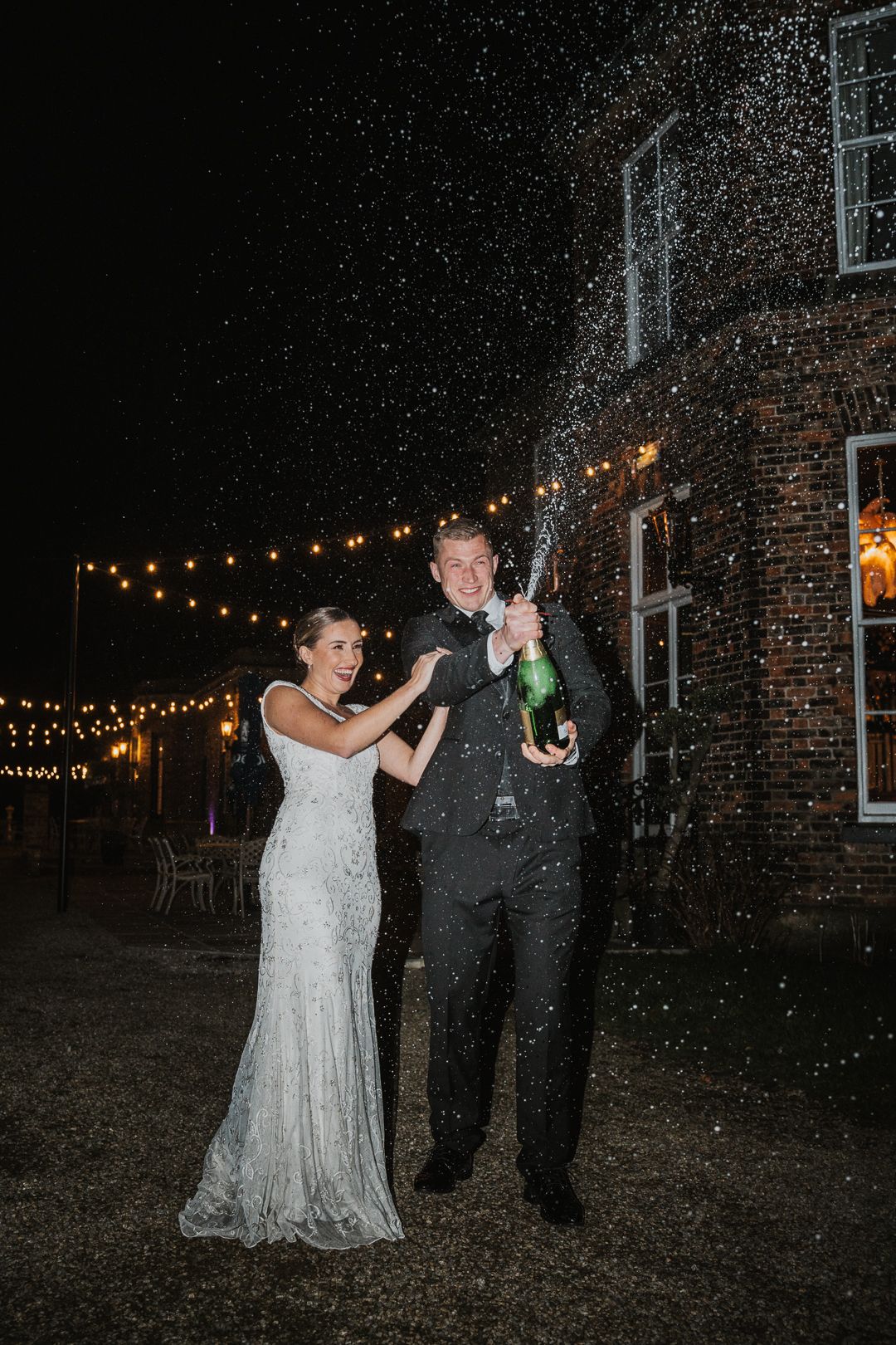 Wedding couple dances outside at night under romantic string lights in the garden while snow gently falls around them.