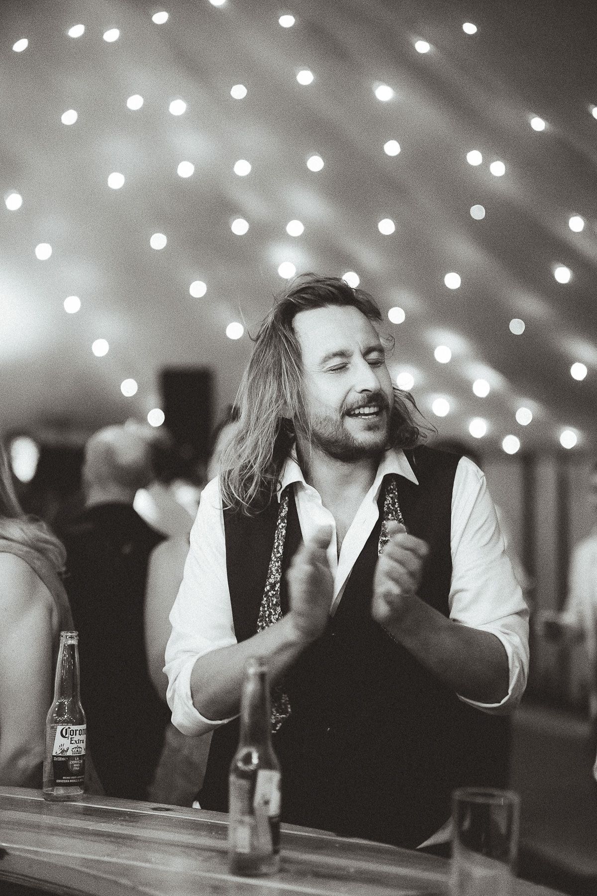 Black and white image shows people gathered at a bar with string lights glowing overhead creating a festive atmosphere.