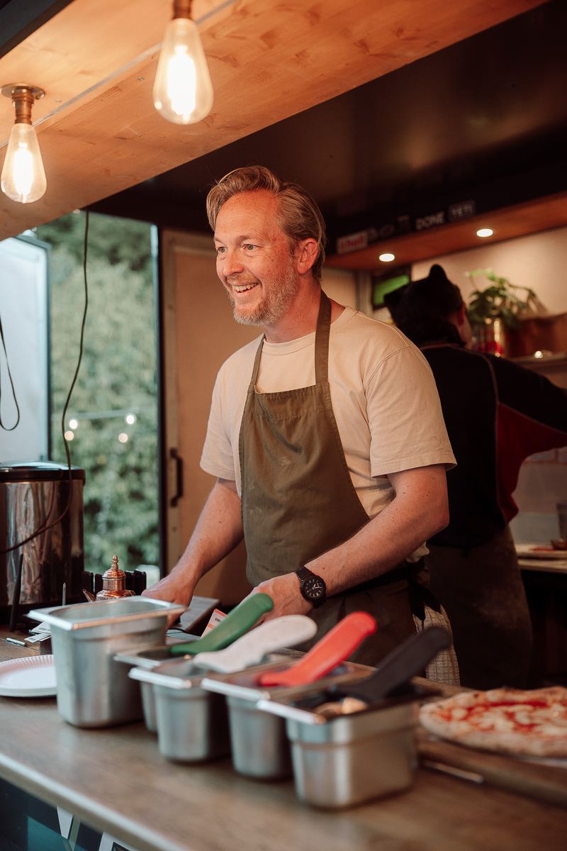 A chef in a brown apron works at a food prep station with metal containers and pendant lights overhead.