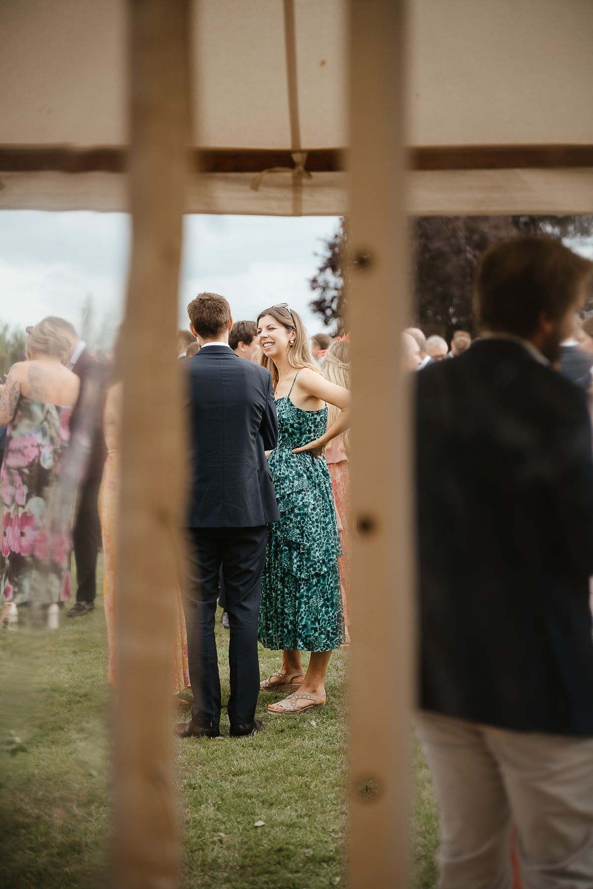 Outdoor event gathering viewed through wooden fence slats showing guests in formal attire including a green floral dress.
