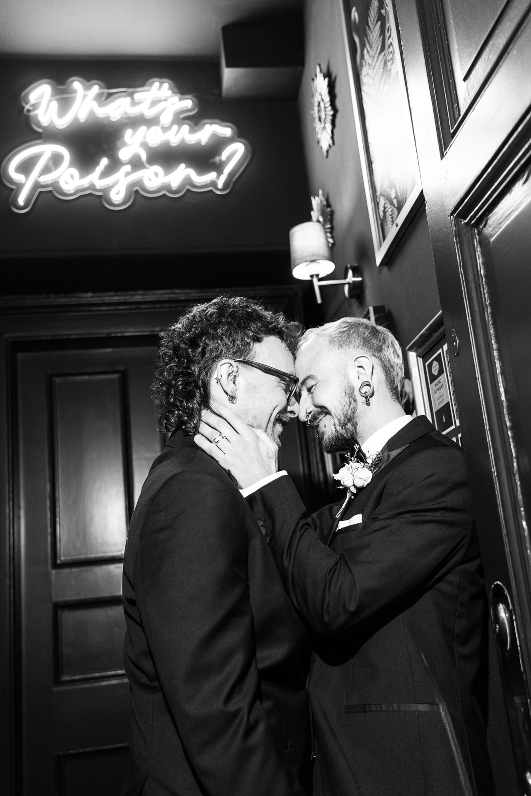 Romantic black and white moment under neon 'Will You Be Mine' sign captures tender intimacy between two people in formal wear.