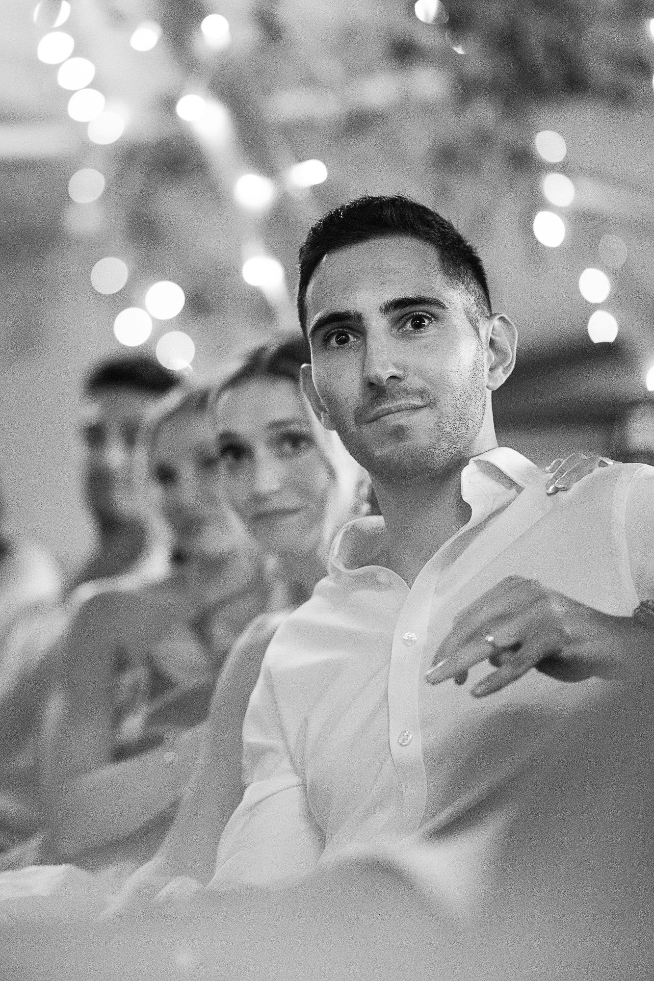 Black and white photo of a man in a white shirt at a wedding reception, with bokeh lights and guests in background.