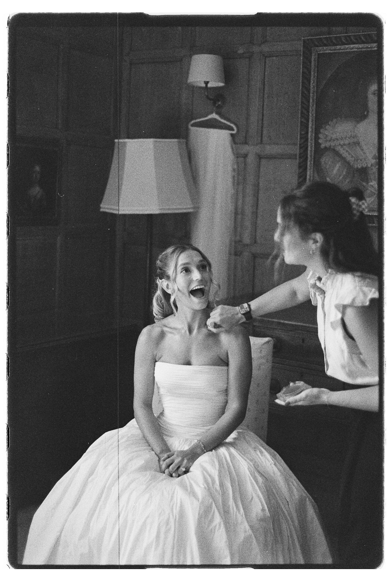 Black and white photo of a bride in a strapless gown reacting with surprise as a woman applies her makeup.