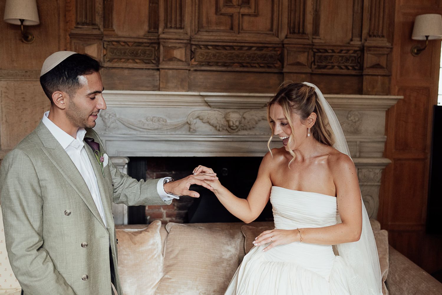 Groom in gray suit placing wedding ring on bride in strapless white gown, smiling by ornate fireplace.