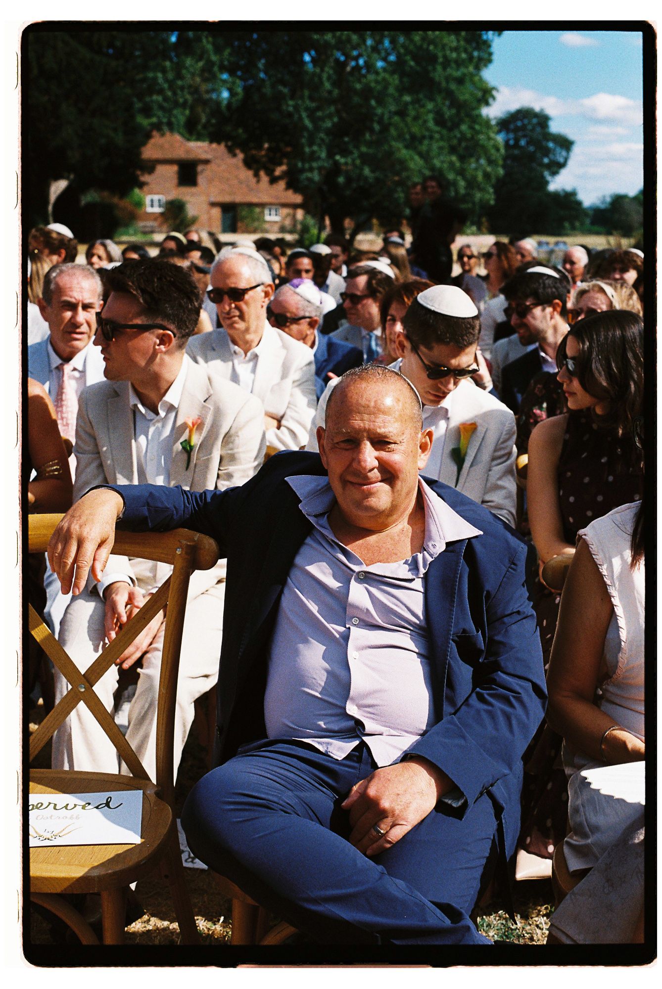 Man in blue suit relaxing at an outdoor wedding ceremony with guests seated on wooden chairs in a sunny garden.