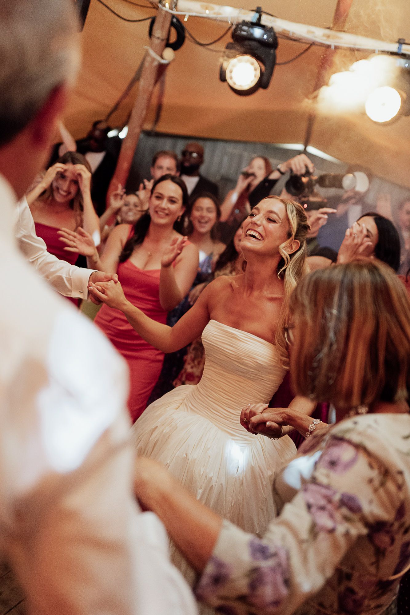 Bride in white strapless gown laughing joyfully while dancing with guests at a lively wedding reception.