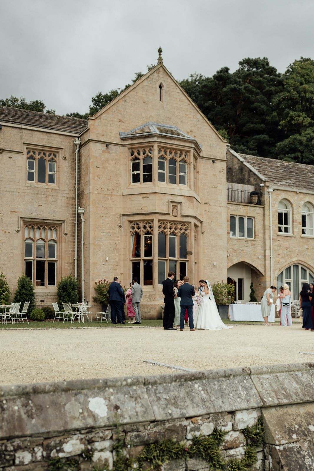 A wedding party gathers outside a grand Gothic stone manor house on a cloudy day.