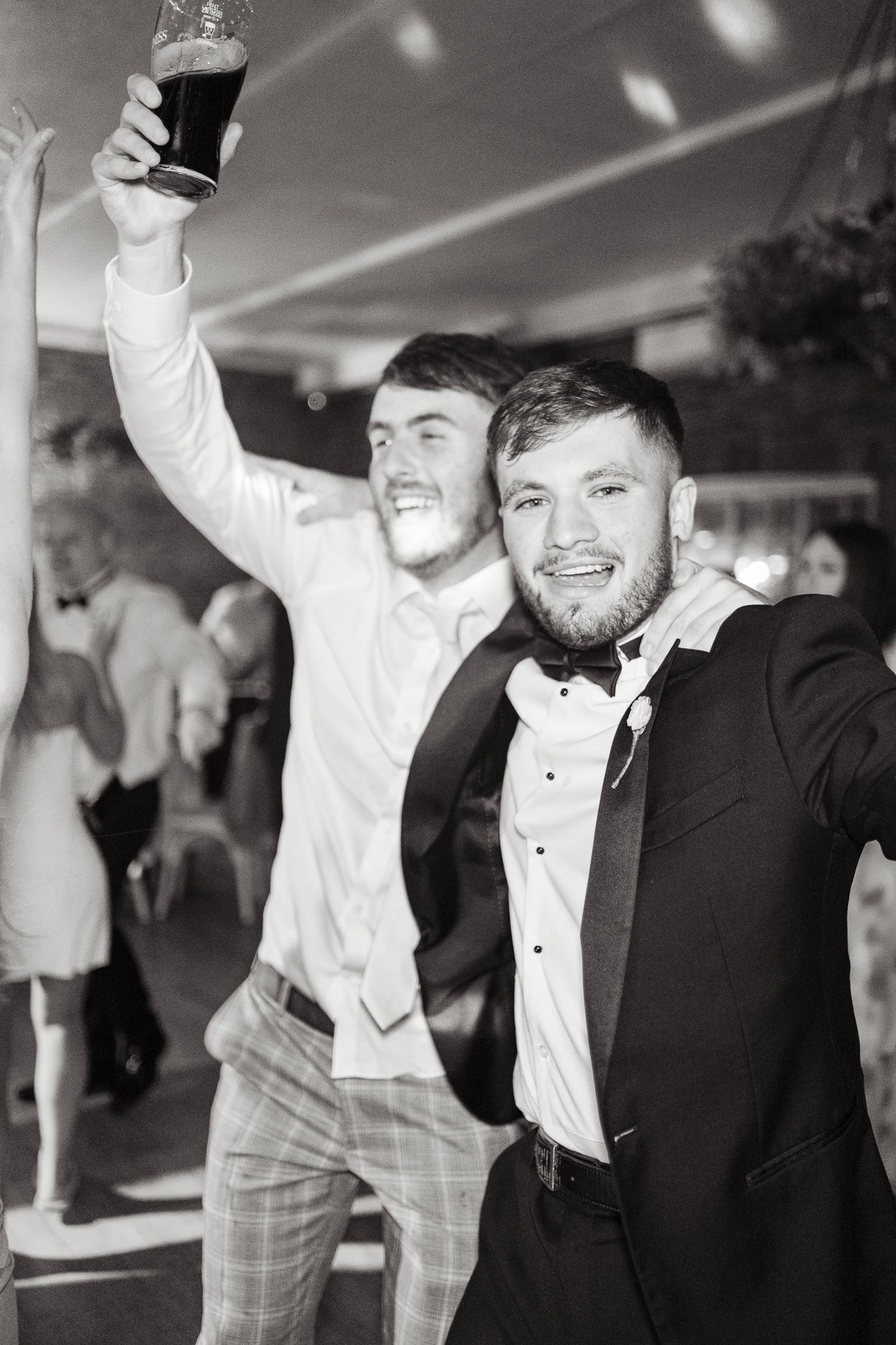 Two friends celebrating at a formal event, with one raising a drink in the air while both smile in black and white photo.