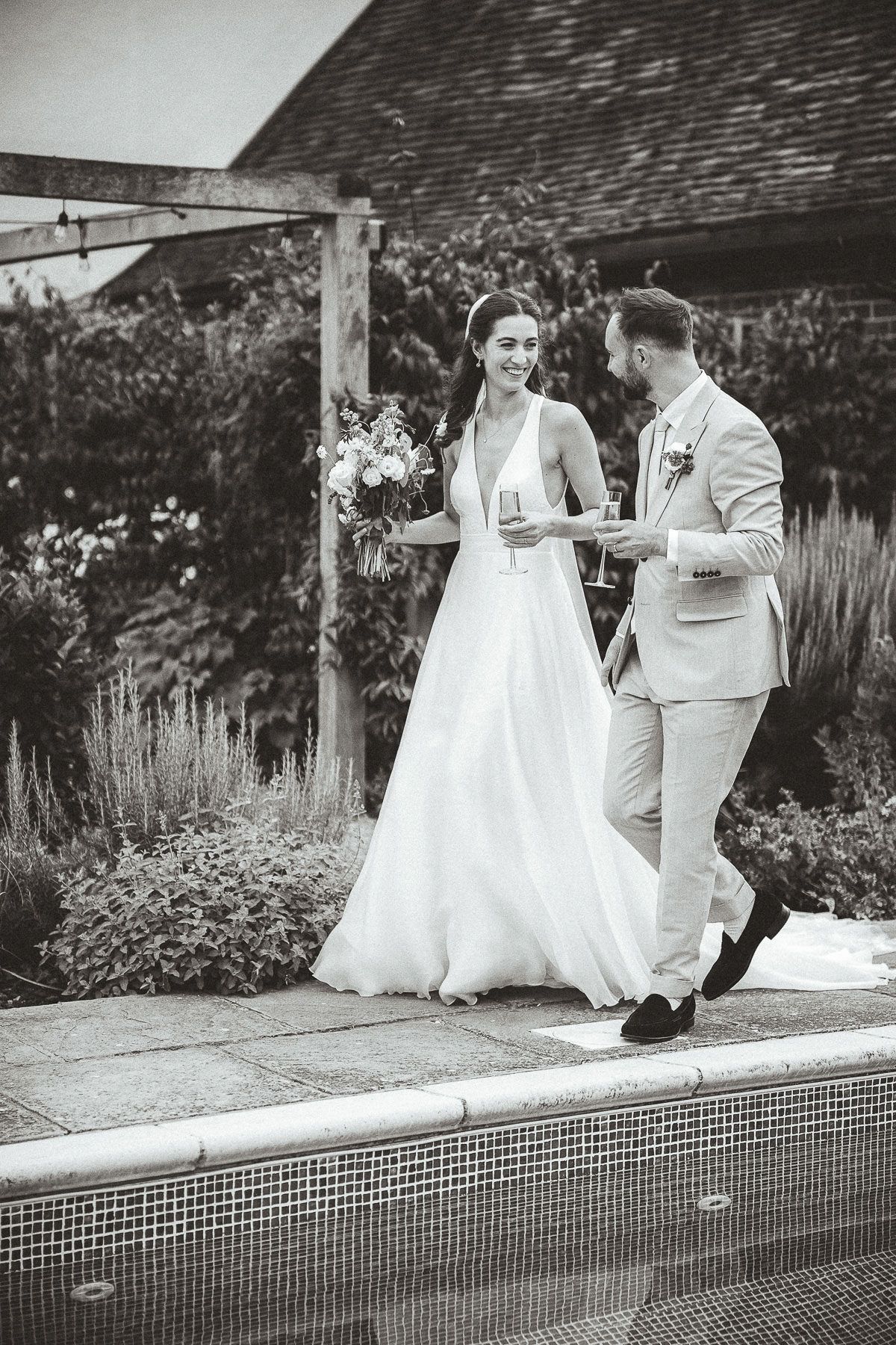 A couple in wedding attire shares a playful moment while sitting on the edge of a swimming pool in a garden setting.