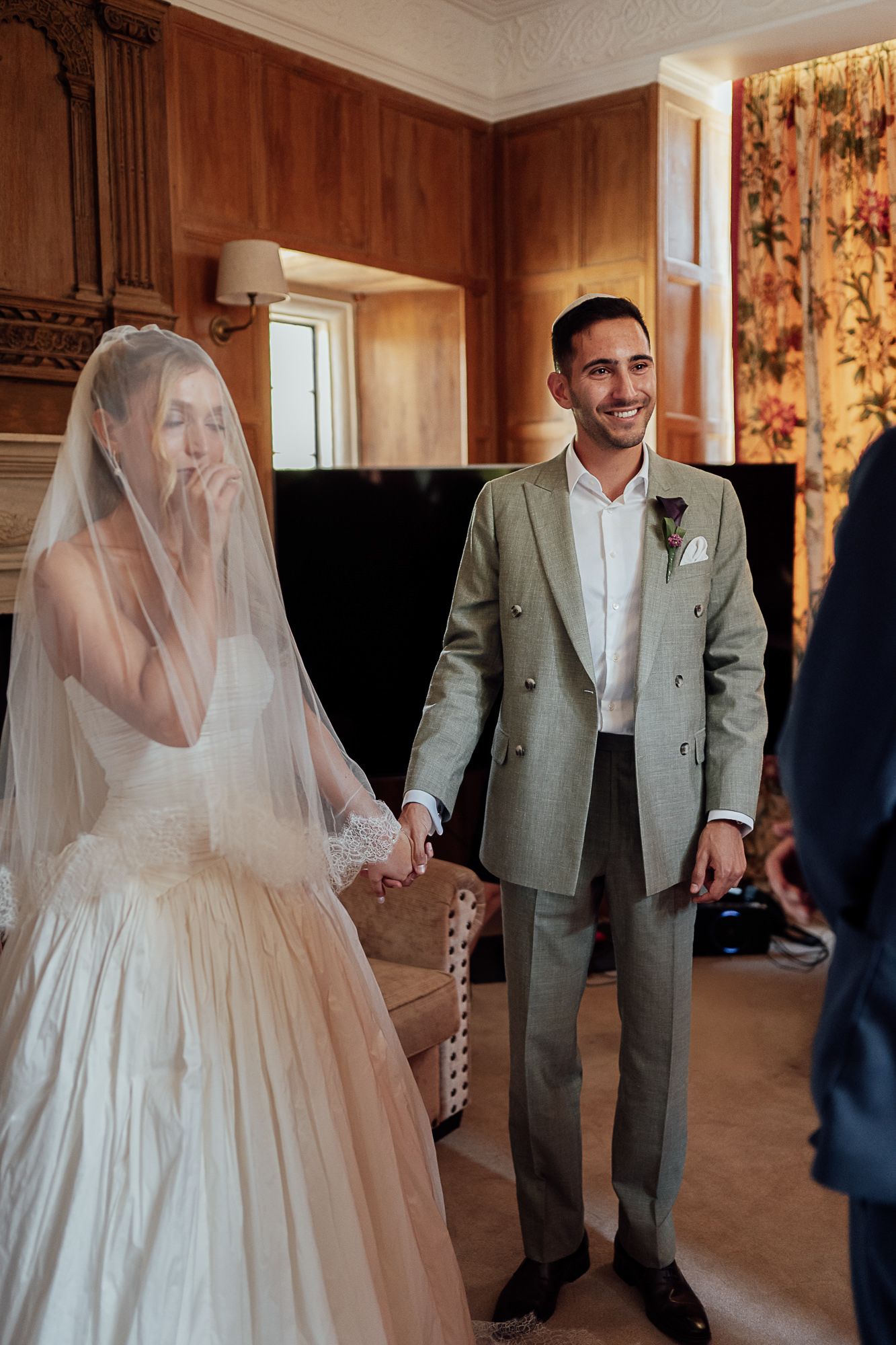 Smiling groom in gray suit holds emotional bride in white wedding dress and veil indoors.
