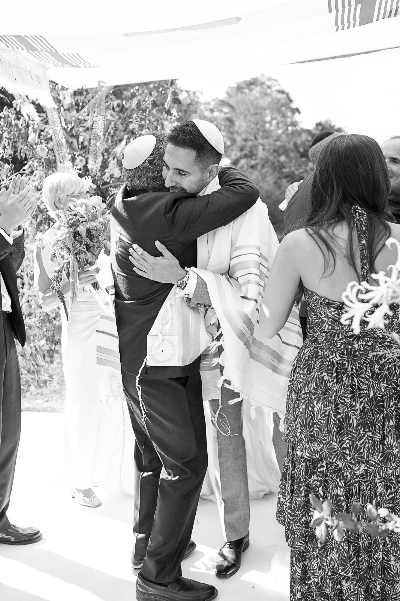 Black and white photo of groom hugging guest at Jewish wedding ceremony outdoors.