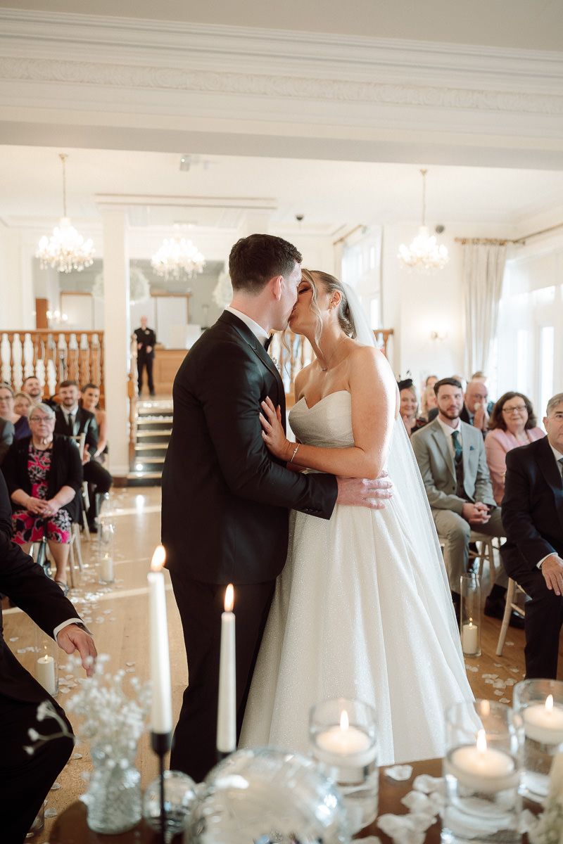 Couple shares their first kiss during an elegant indoor wedding ceremony with crystal chandeliers and candlelit decor.