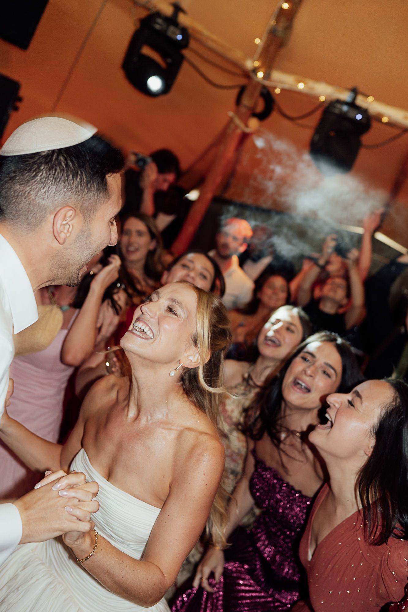 Bride and groom share a joyful dance surrounded by laughing wedding guests at a lively Jewish wedding reception.