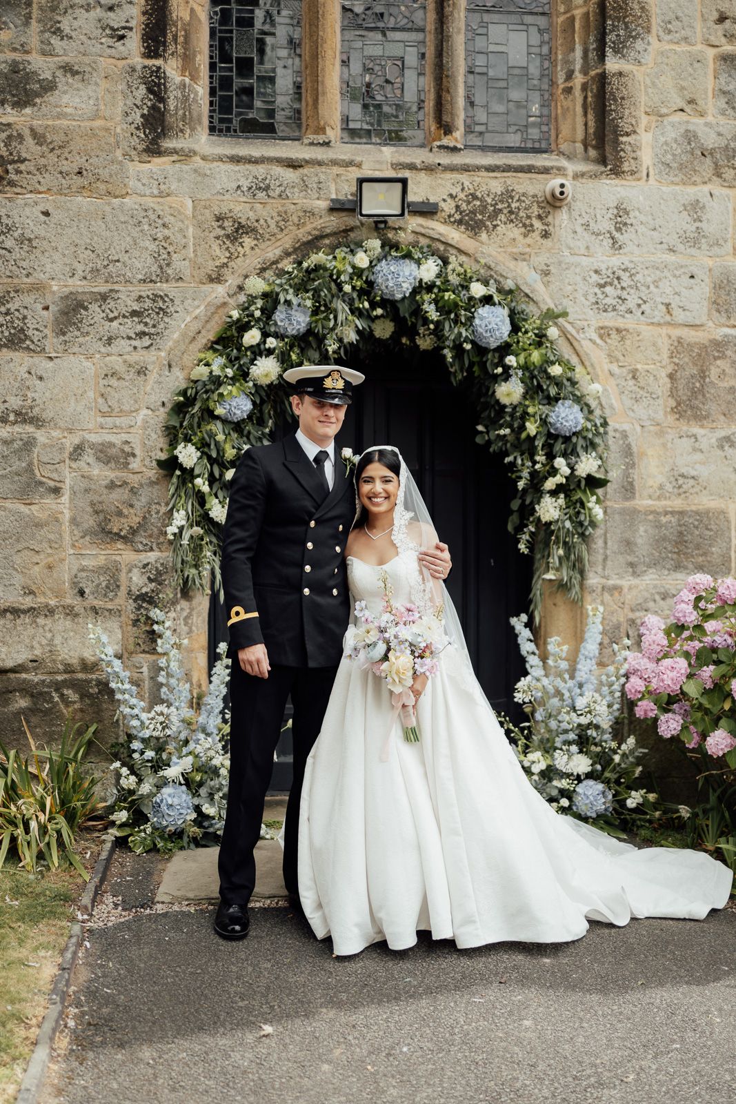 Bride and groom pose in naval uniform before a floral arch at a stone church wedding venue.