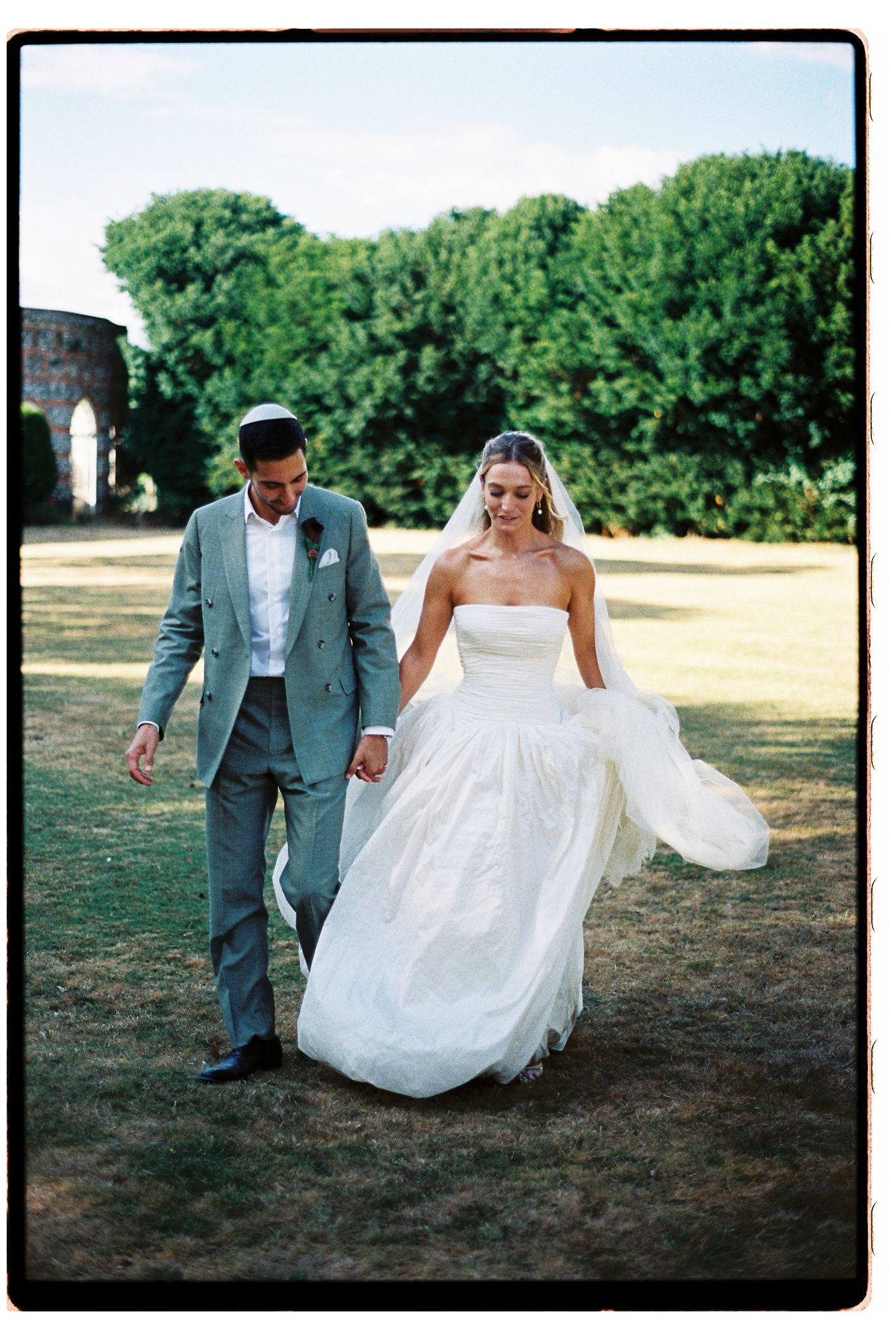 Bride in strapless white gown and groom in gray suit hold hands walking outdoors at elegant garden wedding venue.