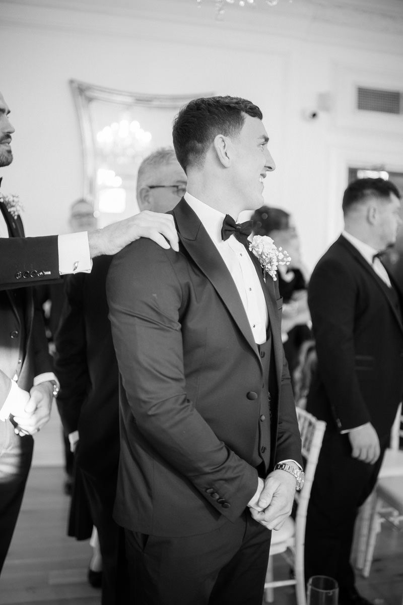 Black and white photo of a groom in a tuxedo and bowtie at an elegant wedding reception.
