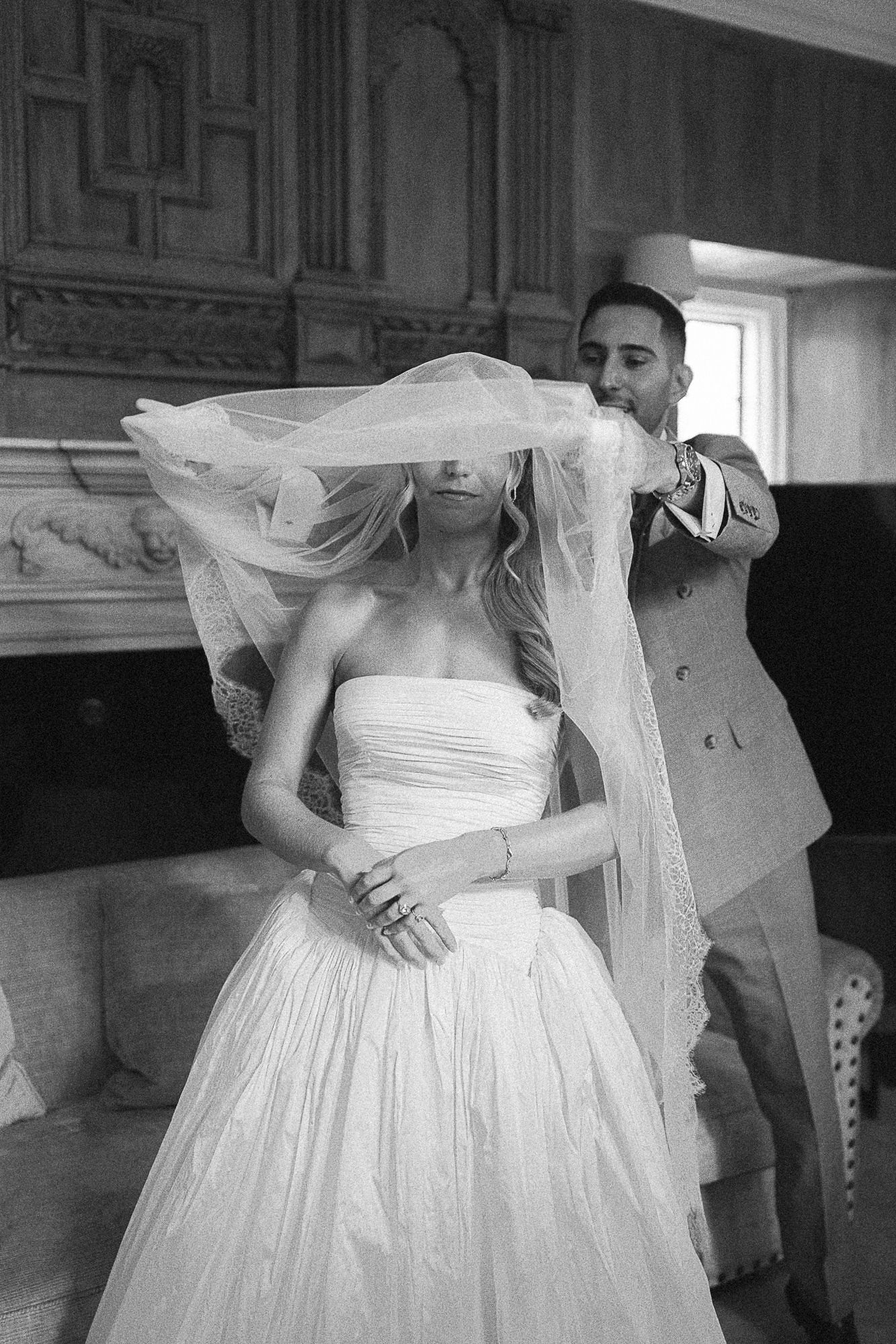 Black and white photo of groom lifting bride's veil indoors during wedding preparations.