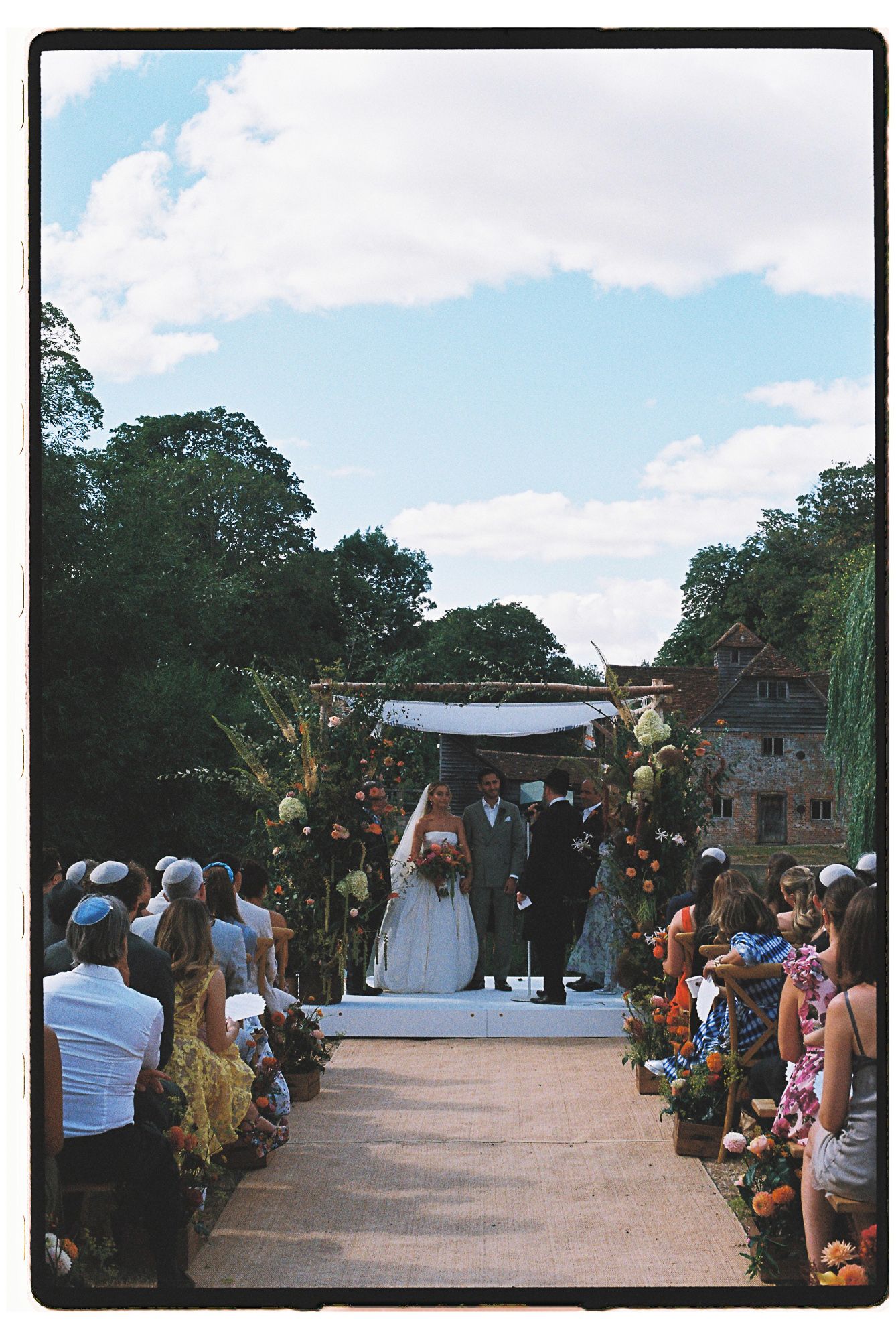 Bride and groom walk down outdoor aisle at garden wedding ceremony with floral decorations and guests.