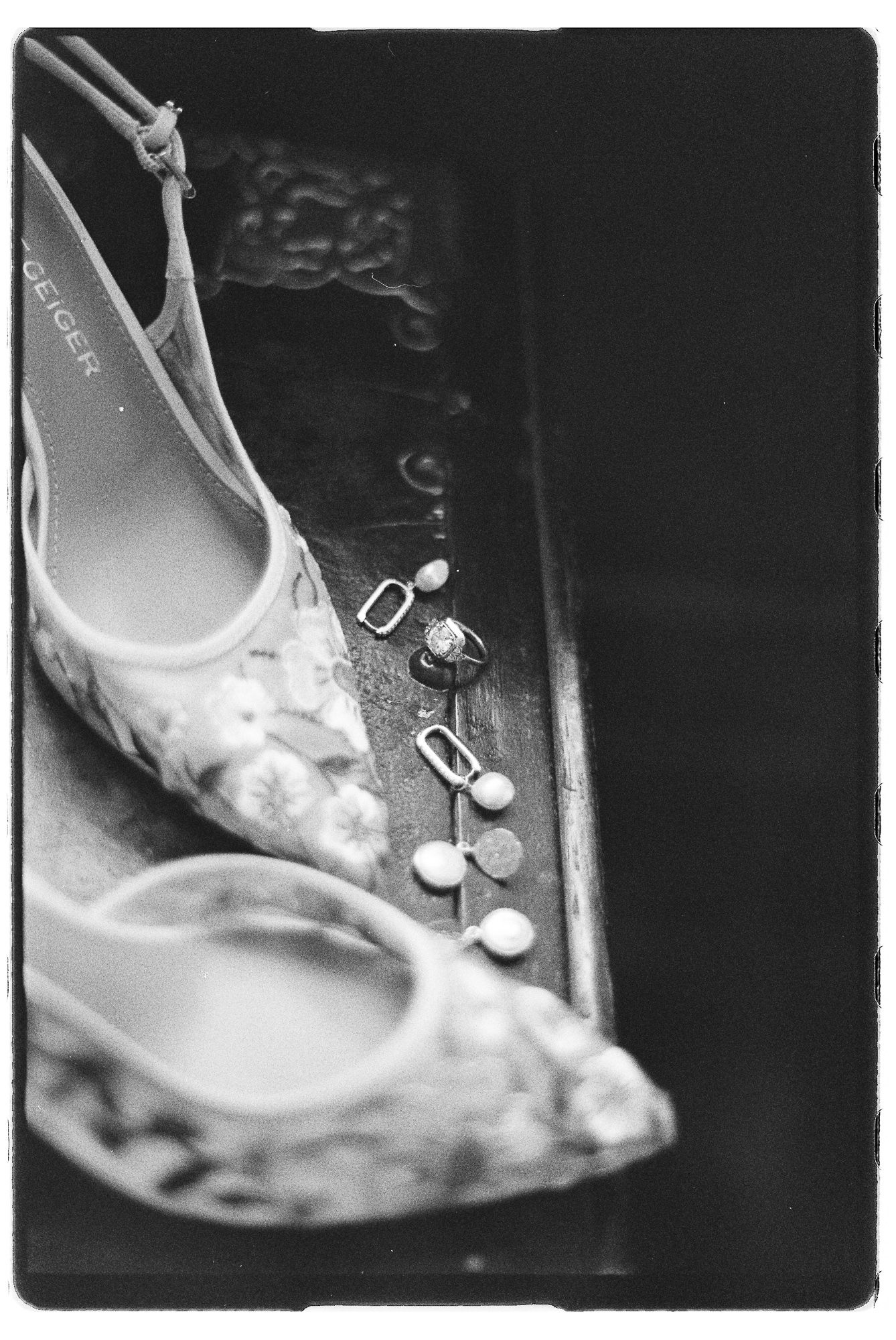 Black and white photo of floral bridal heels beside delicate pearl earrings and rings on a wooden surface.