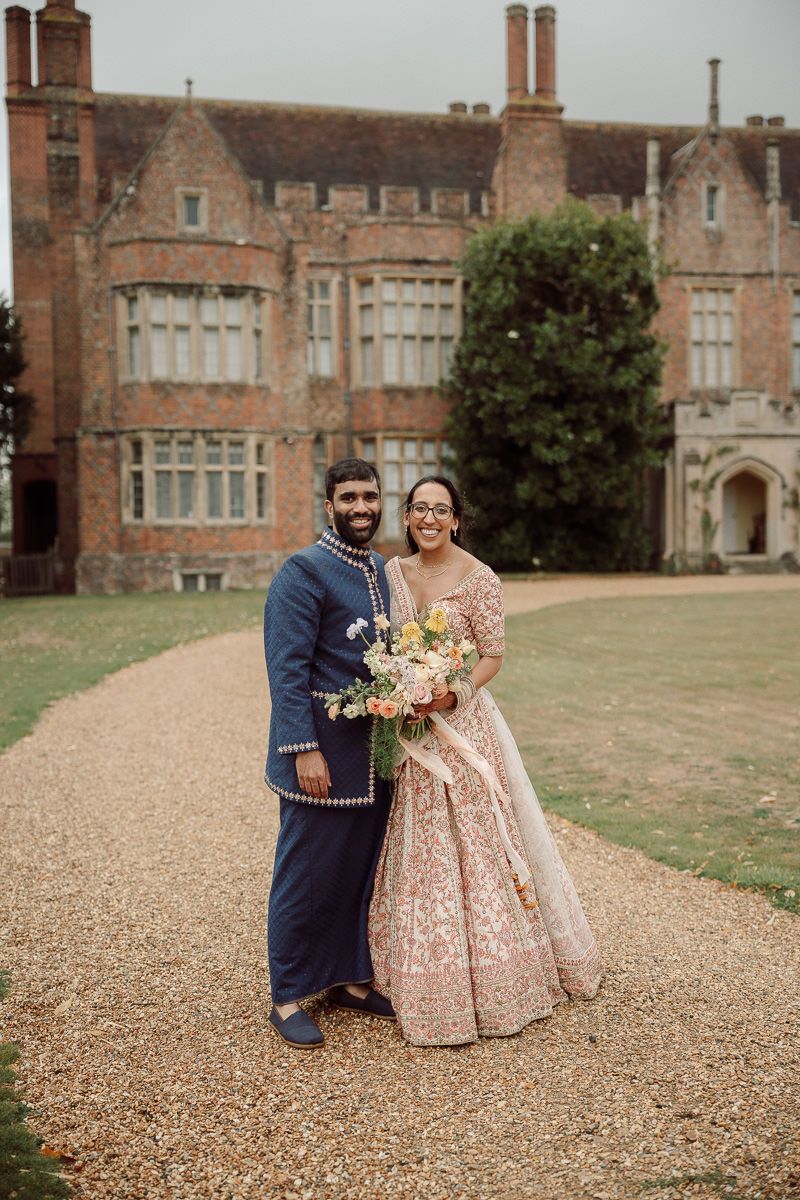 Couple in wedding attire posing on gravel path in front of historic mansion.