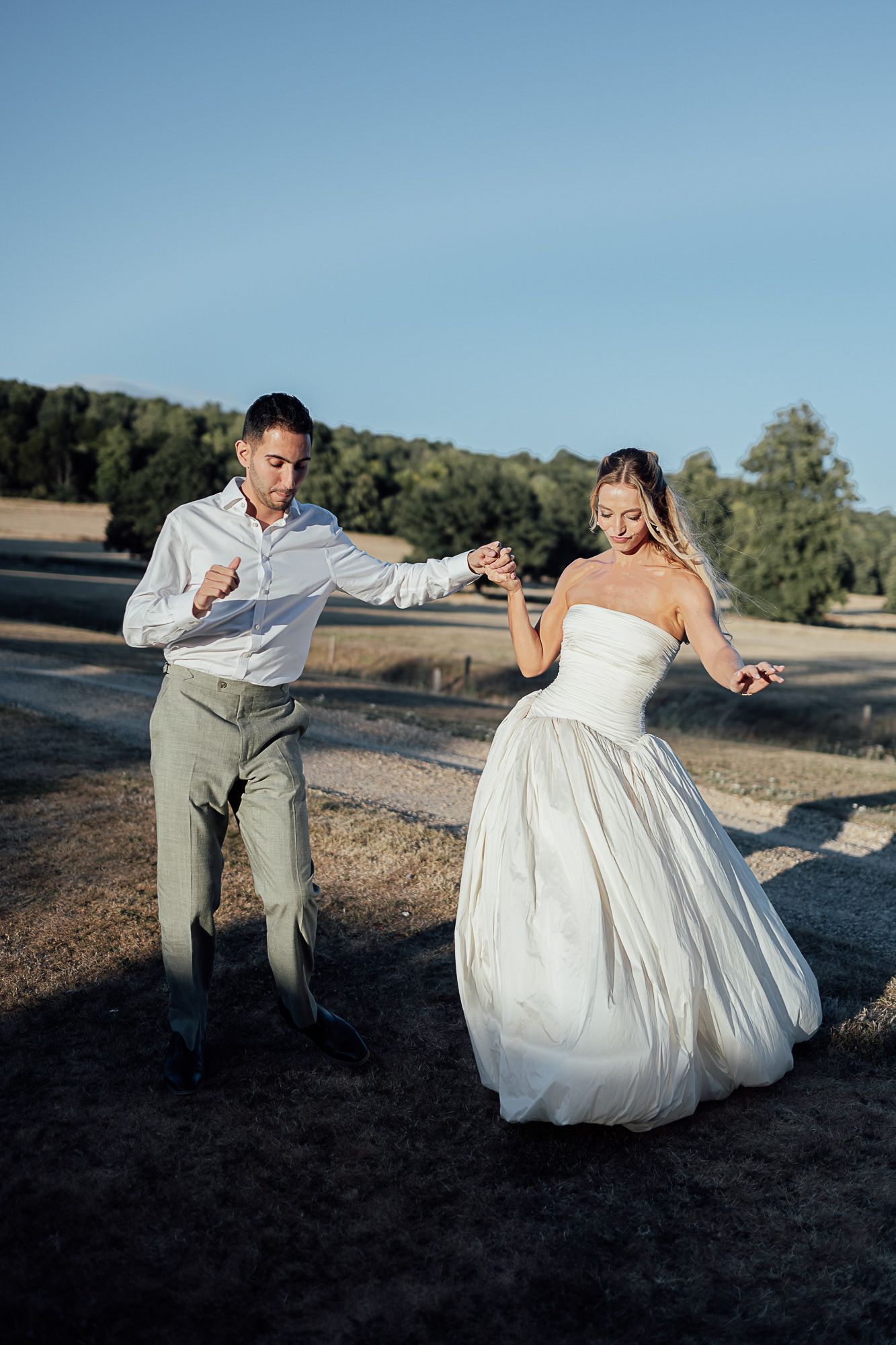 Bride in white strapless gown and groom in gray suit dancing outdoors on sunny day with trees in background.
