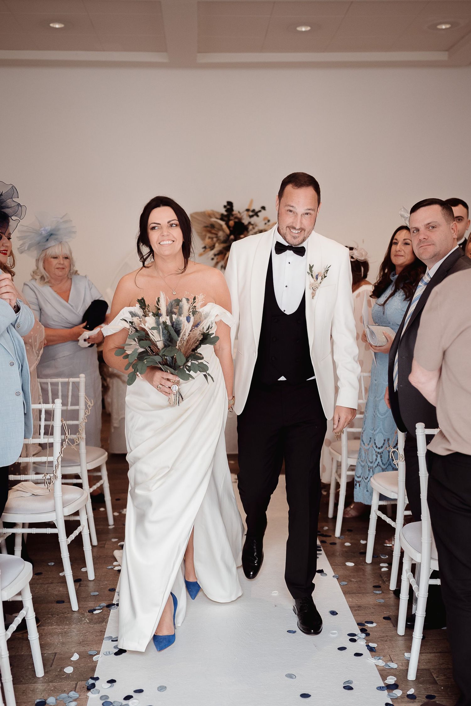 Bride in elegant white gown with blue heels walks down aisle with groom in white dinner jacket during indoor ceremony.