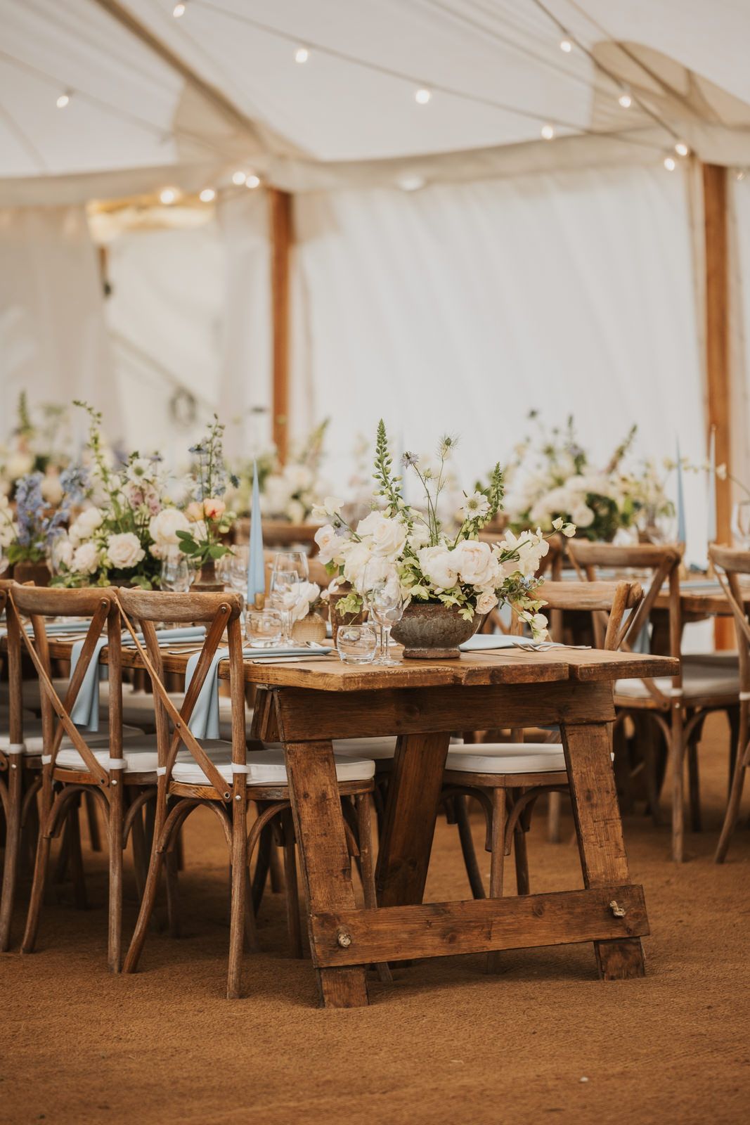 Rustic wooden farm tables decorated with elegant floral centerpieces and place settings in a light-filled reception venue.