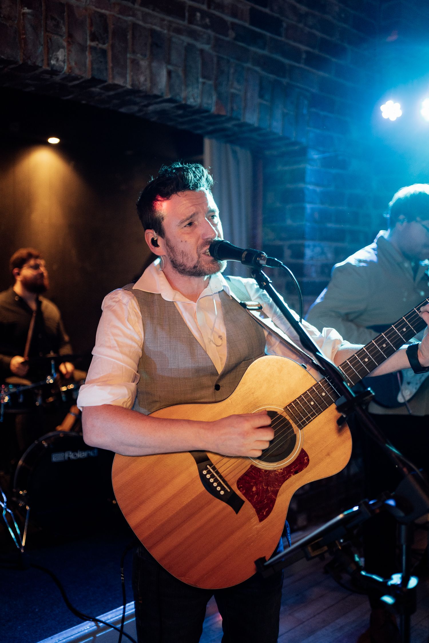 Musician performs with acoustic guitar at intimate venue with blue stage lighting and brick wall backdrop.