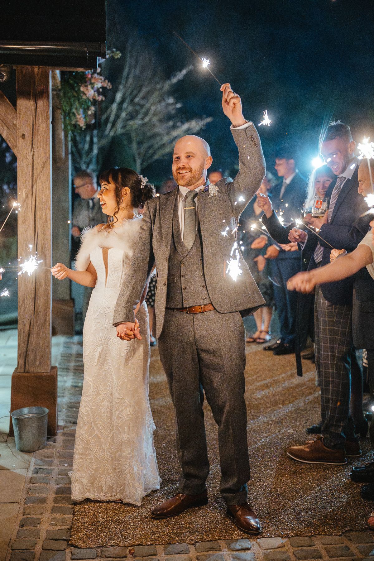 A wedding couple dances under sparklers held by guests at a rustic outdoor nighttime celebration.