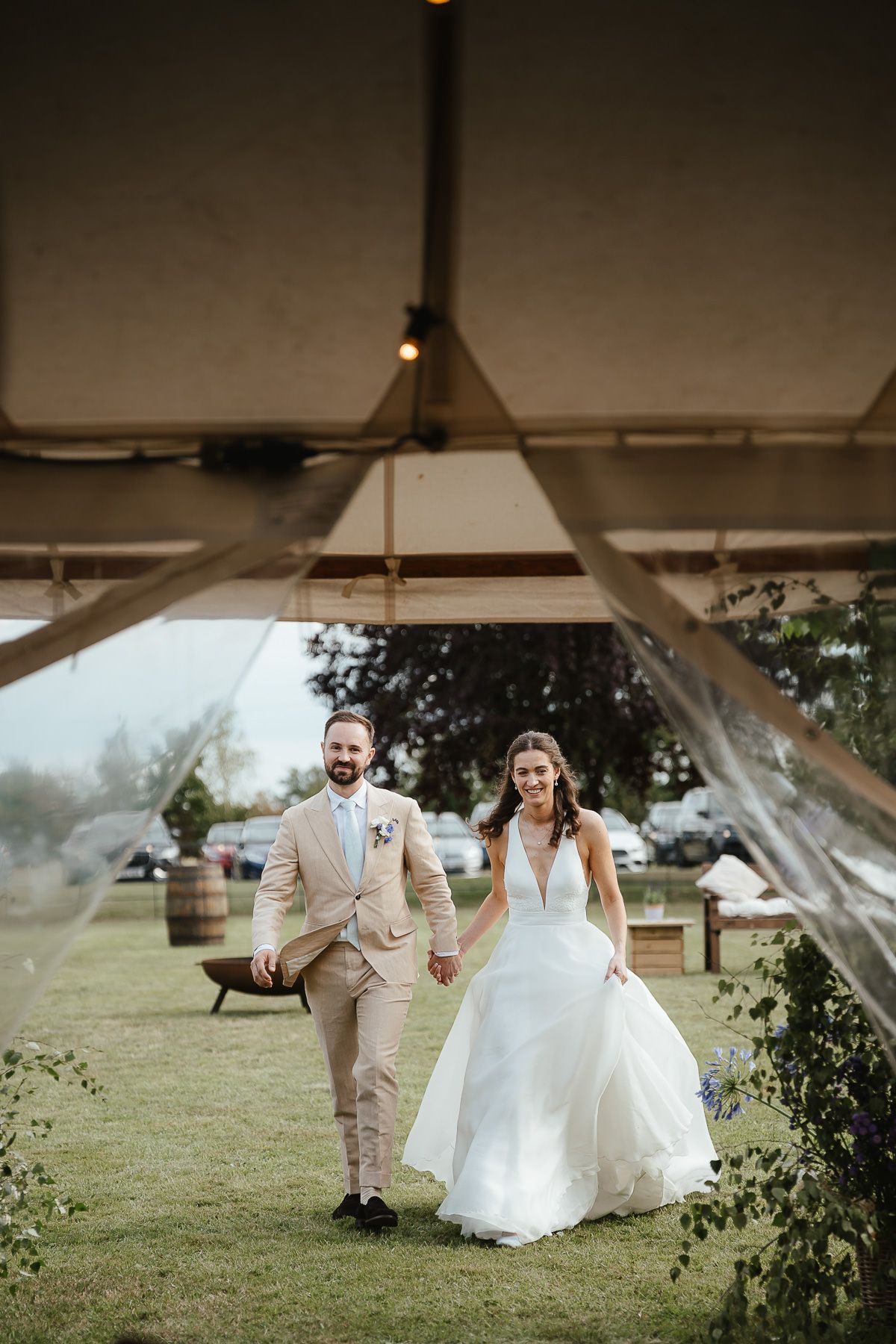 A bride and groom hold hands walking under a white tent during their outdoor wedding celebration.