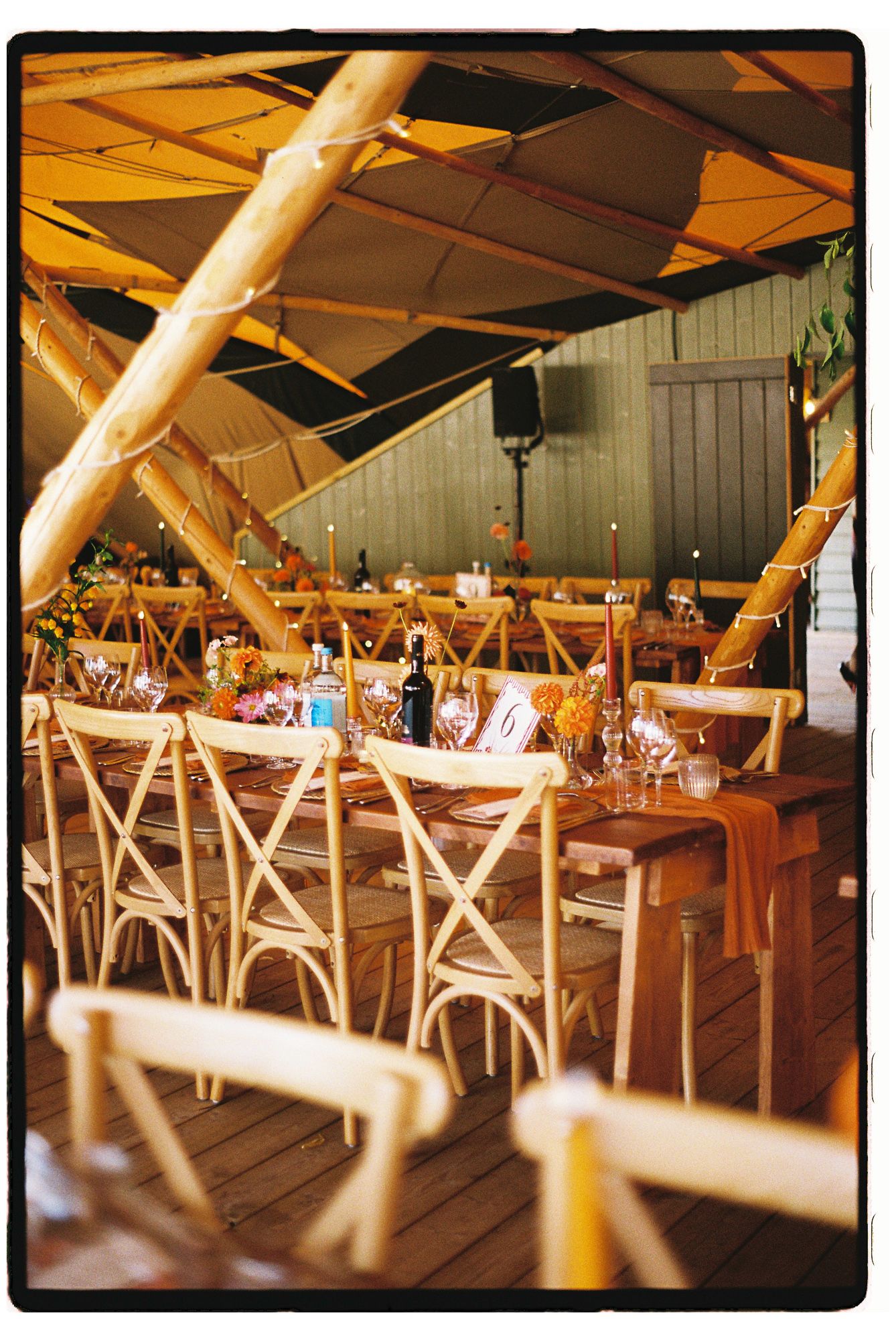 Rustic wedding reception under a tipi tent with wooden cross-back chairs and warm amber decor.