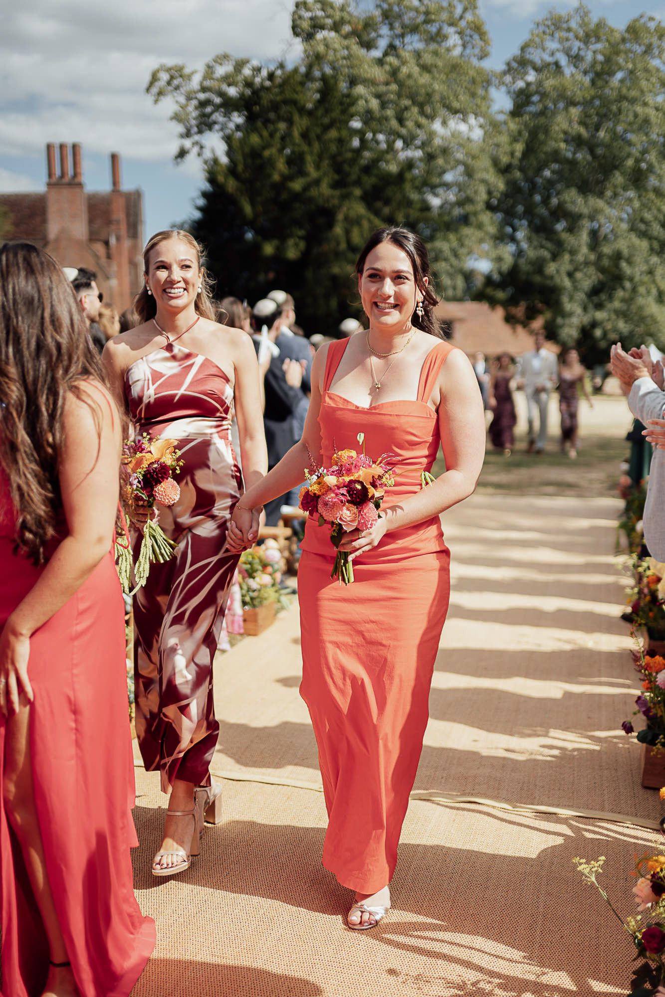 Bridesmaids in coral and floral dresses walk down an outdoor aisle holding colorful bouquets at a sunny wedding.
