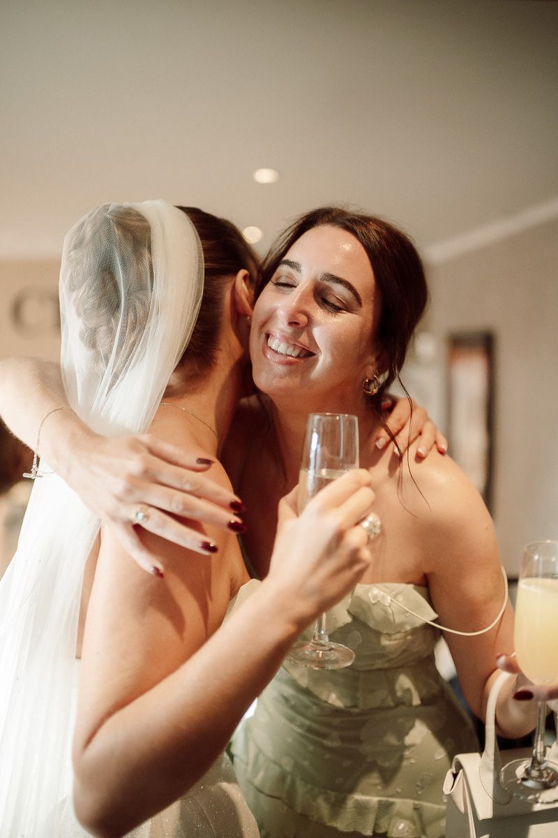 Two women in formal attire share a joyful moment while holding wine glasses at an elegant indoor event.