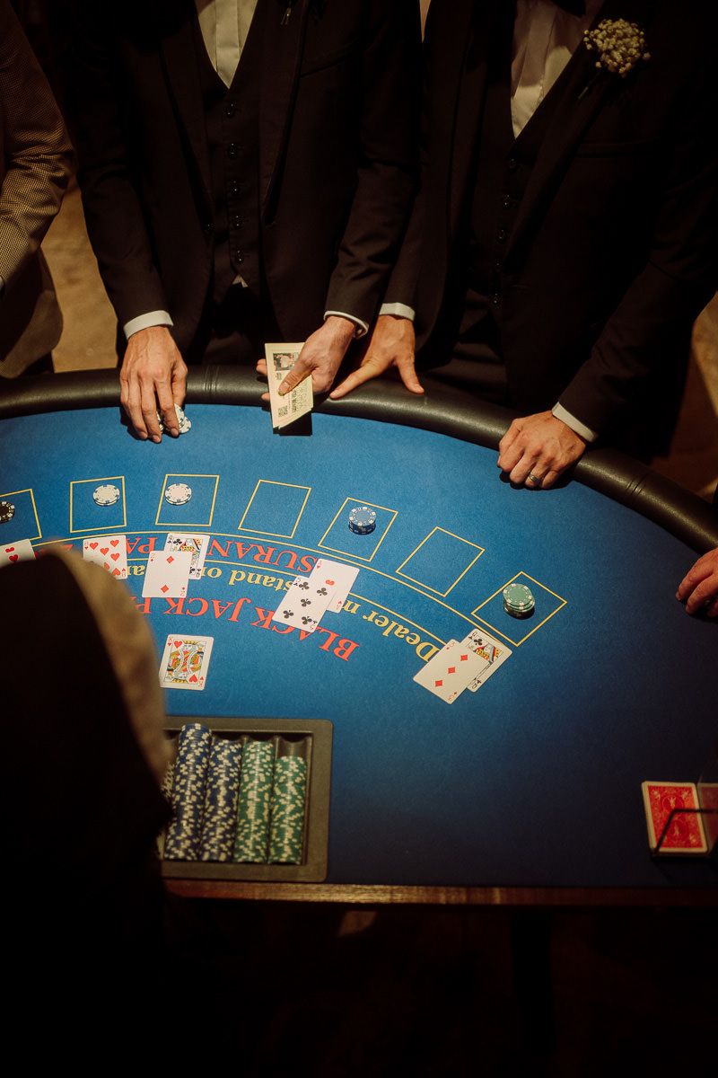 Players in dark suits gather around a blue casino blackjack table with cards and poker chips during a high-stakes game.