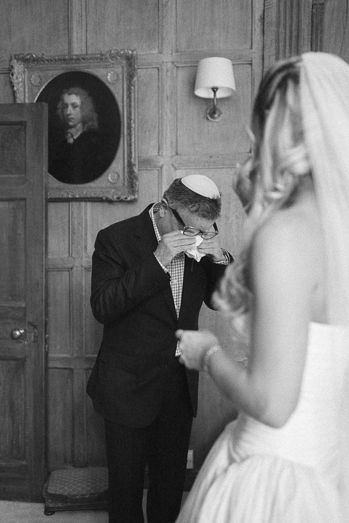 Emotional father wipes tears seeing bride in wedding dress during touching first look moment, black and white photo.