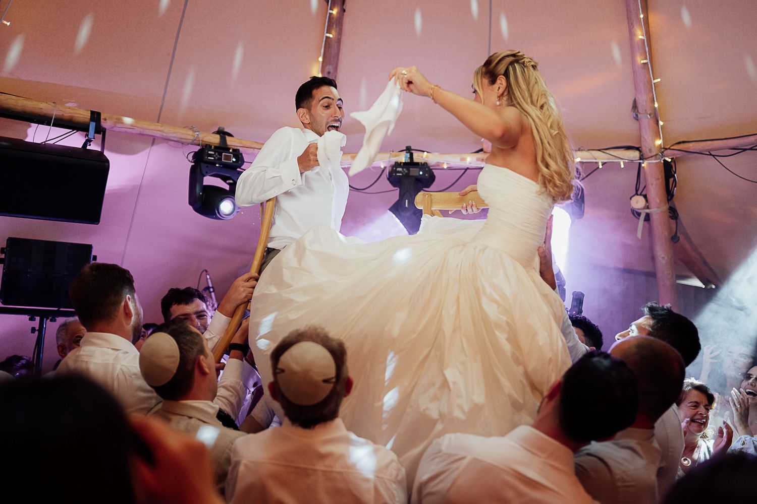 Bride and groom lifted by guests during joyful Jewish wedding hora dance celebration under festive tent lighting.