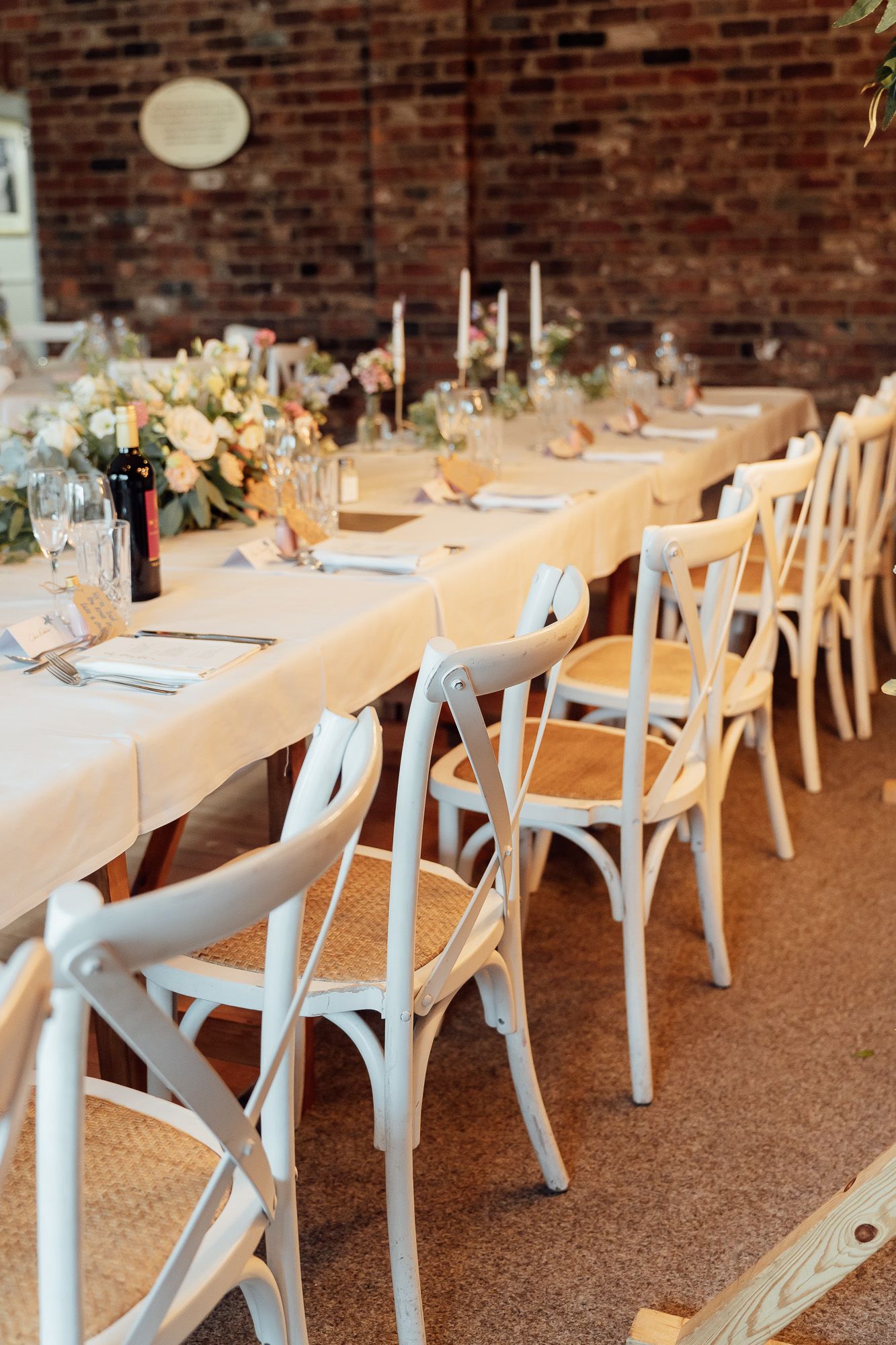 Elegant rustic wedding table setup with white crossback chairs and peach floral centerpieces against exposed brick wall.