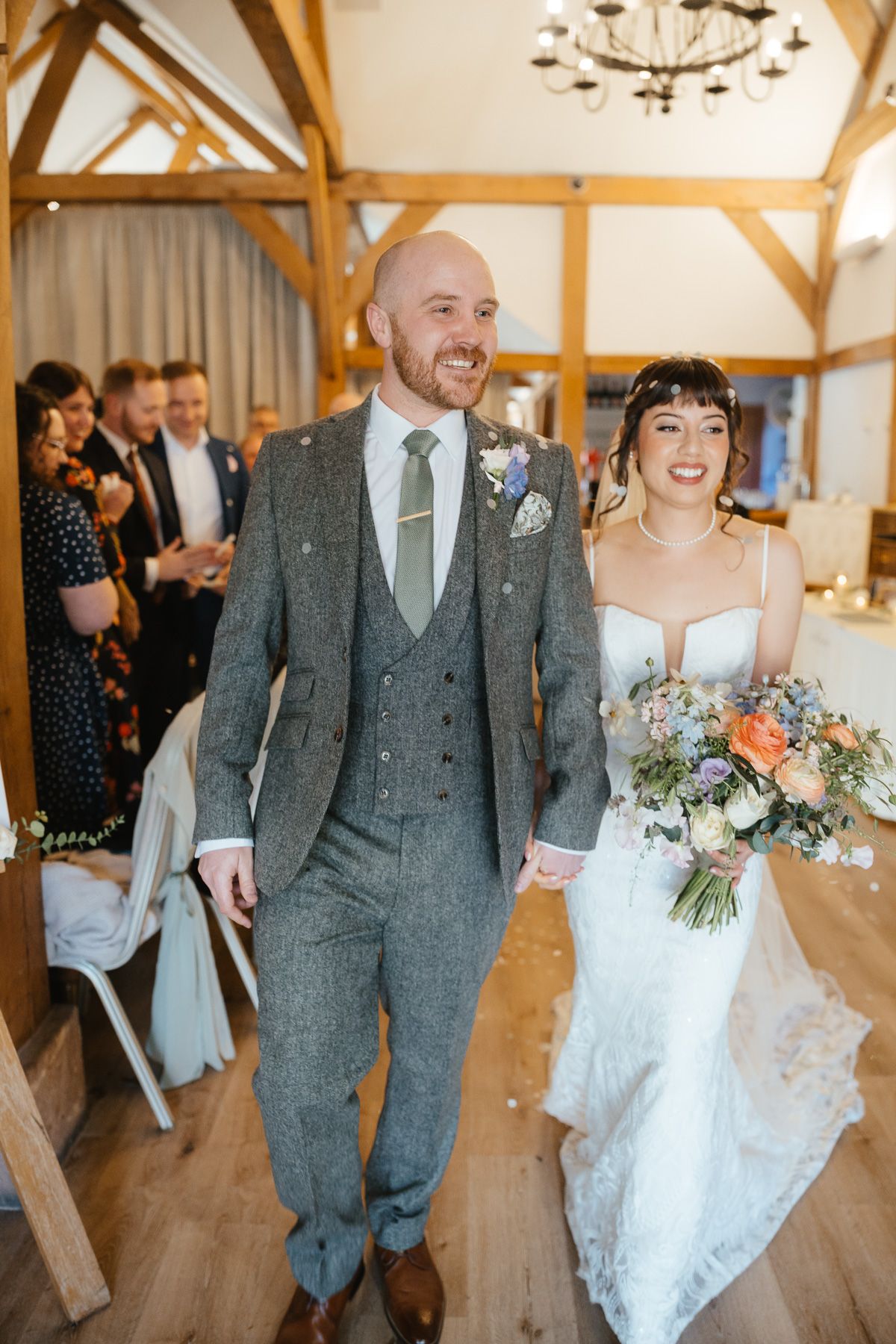 Newlywed couple walks down wooden aisle in rustic venue with bride holding pastel bouquet and groom in grey suit.