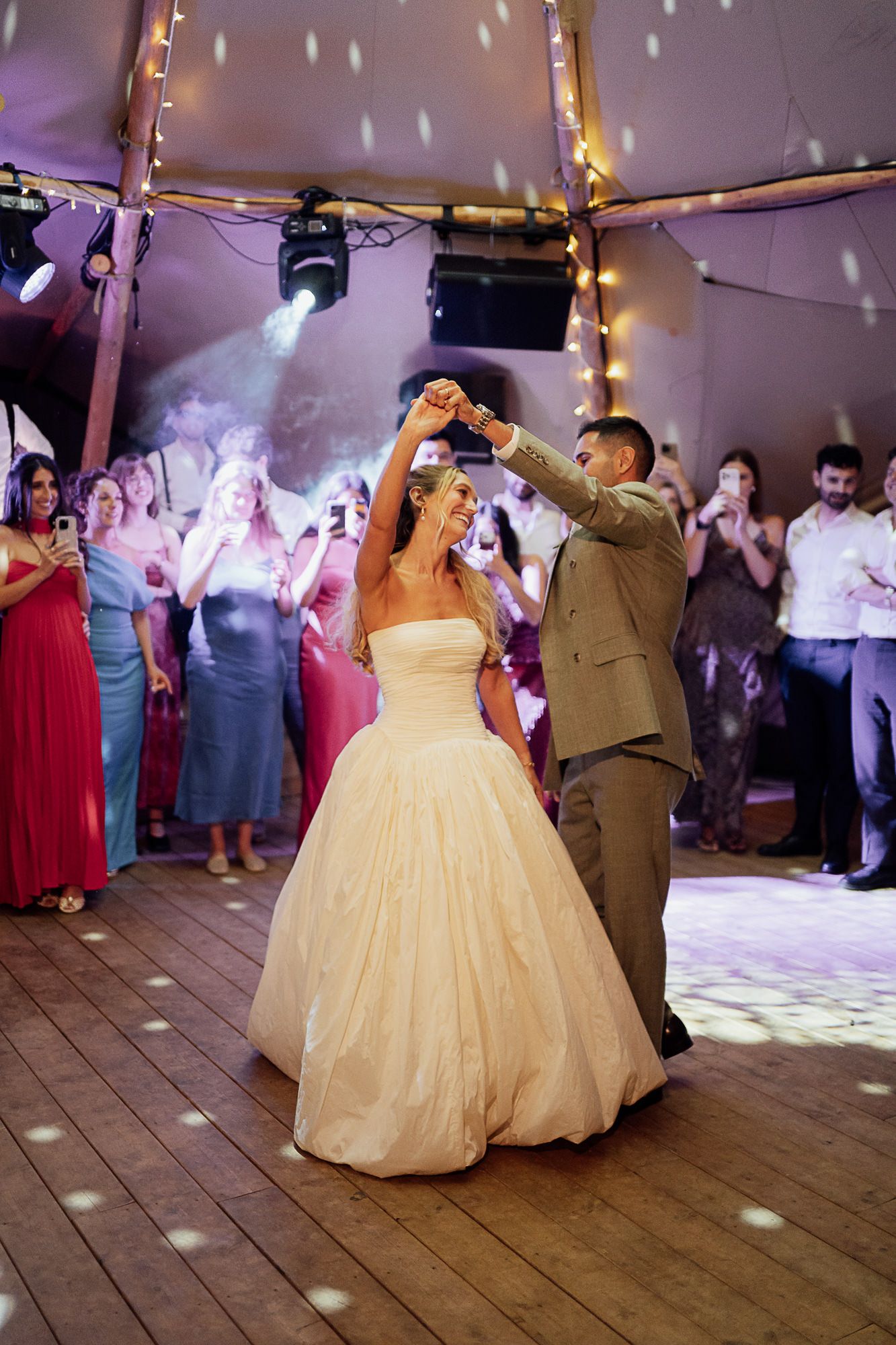 Bride and groom share first dance in a marquee wedding venue with fairy lights and guests watching.