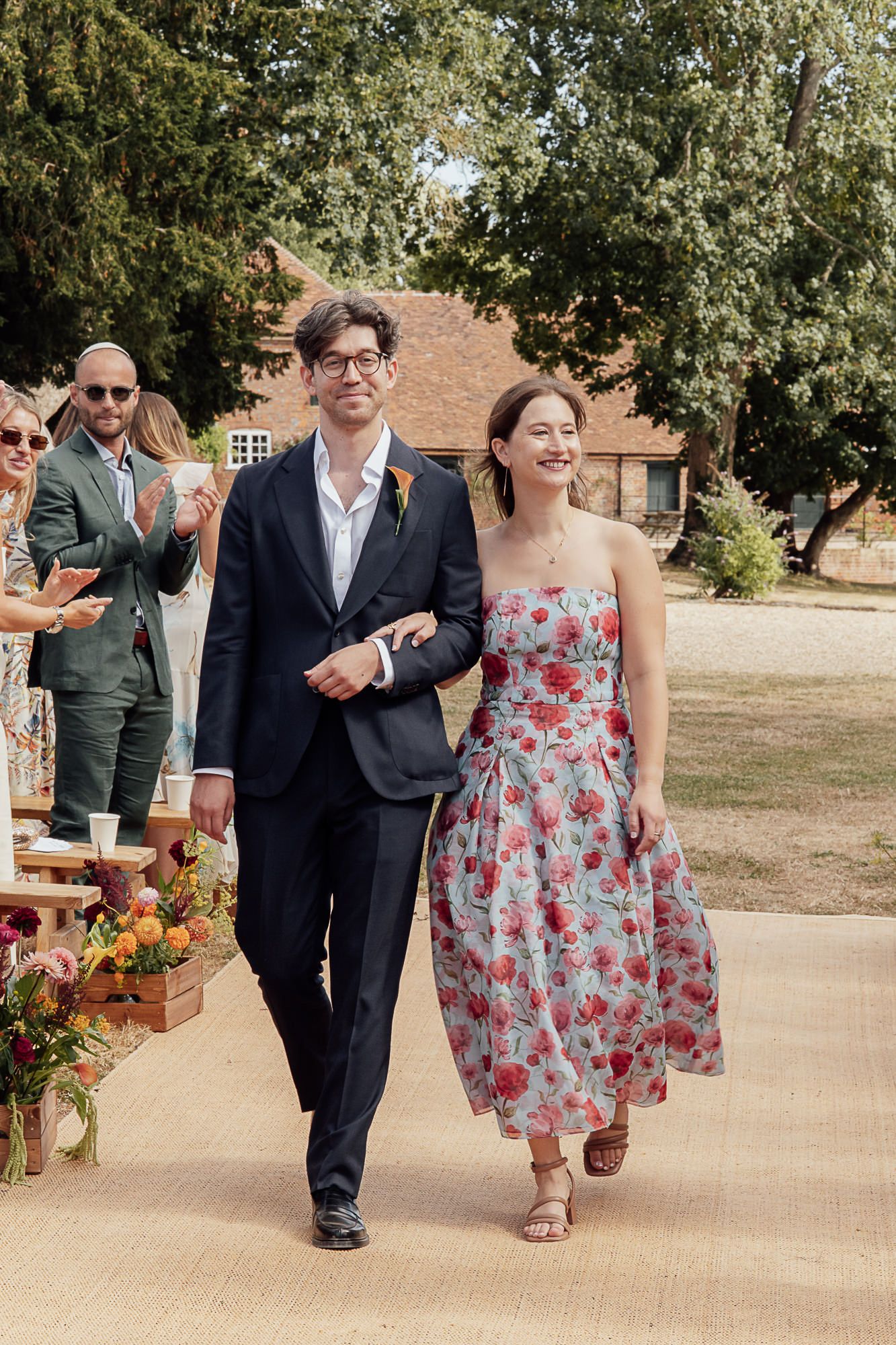 Couple walks down outdoor wedding aisle, groom in navy suit, bride in floral dress, guests applauding, rustic venue.