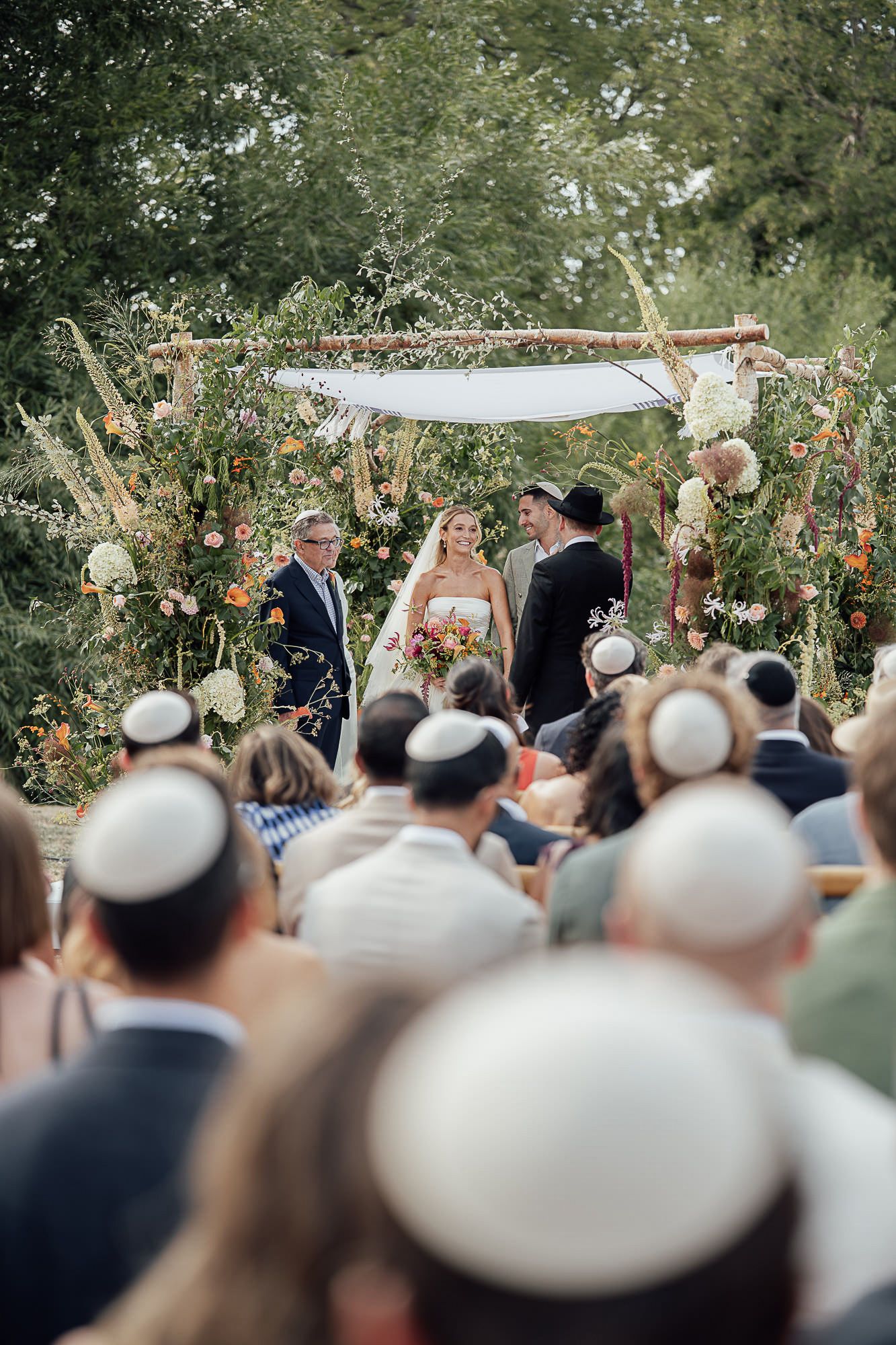 Outdoor Jewish wedding ceremony under a floral chuppa with guests wearing kippahs seated in the foreground.
