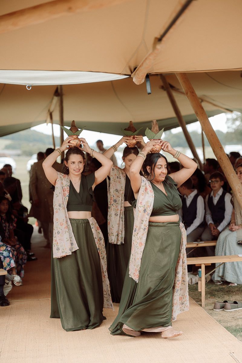 Dancers in olive green dresses and floral scarves perform a synchronized dance routine under a beige tent.