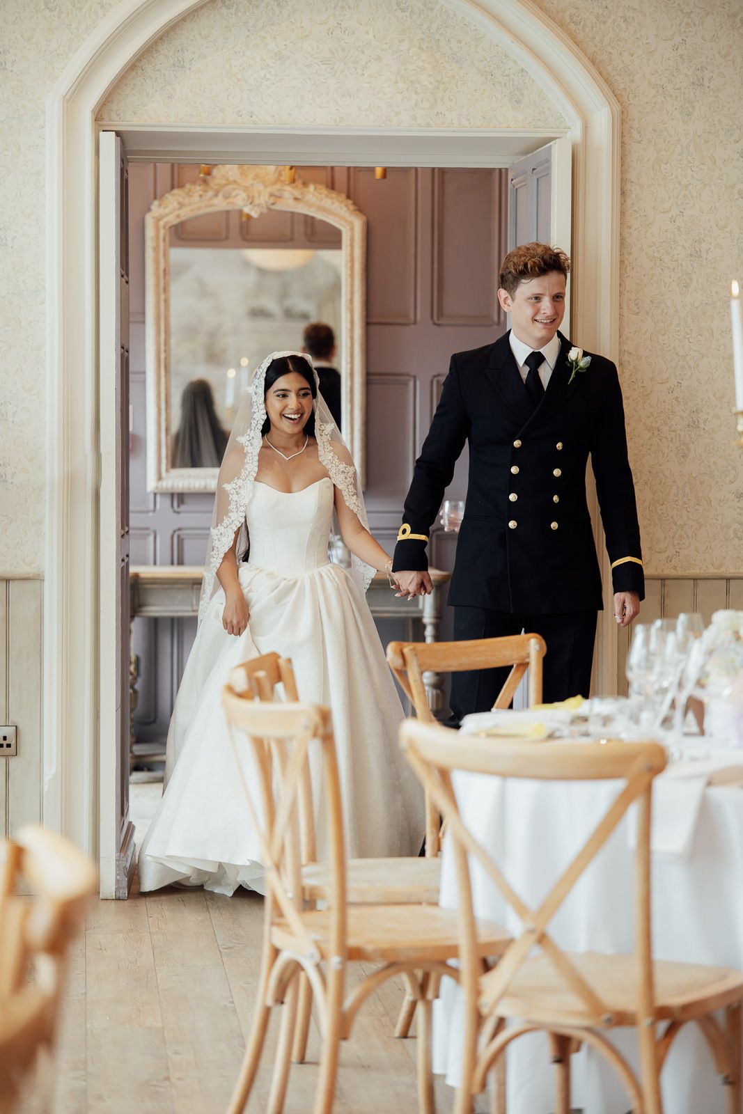 Bride in white gown and groom in navy military suit hold hands entering elegant wedding reception venue.