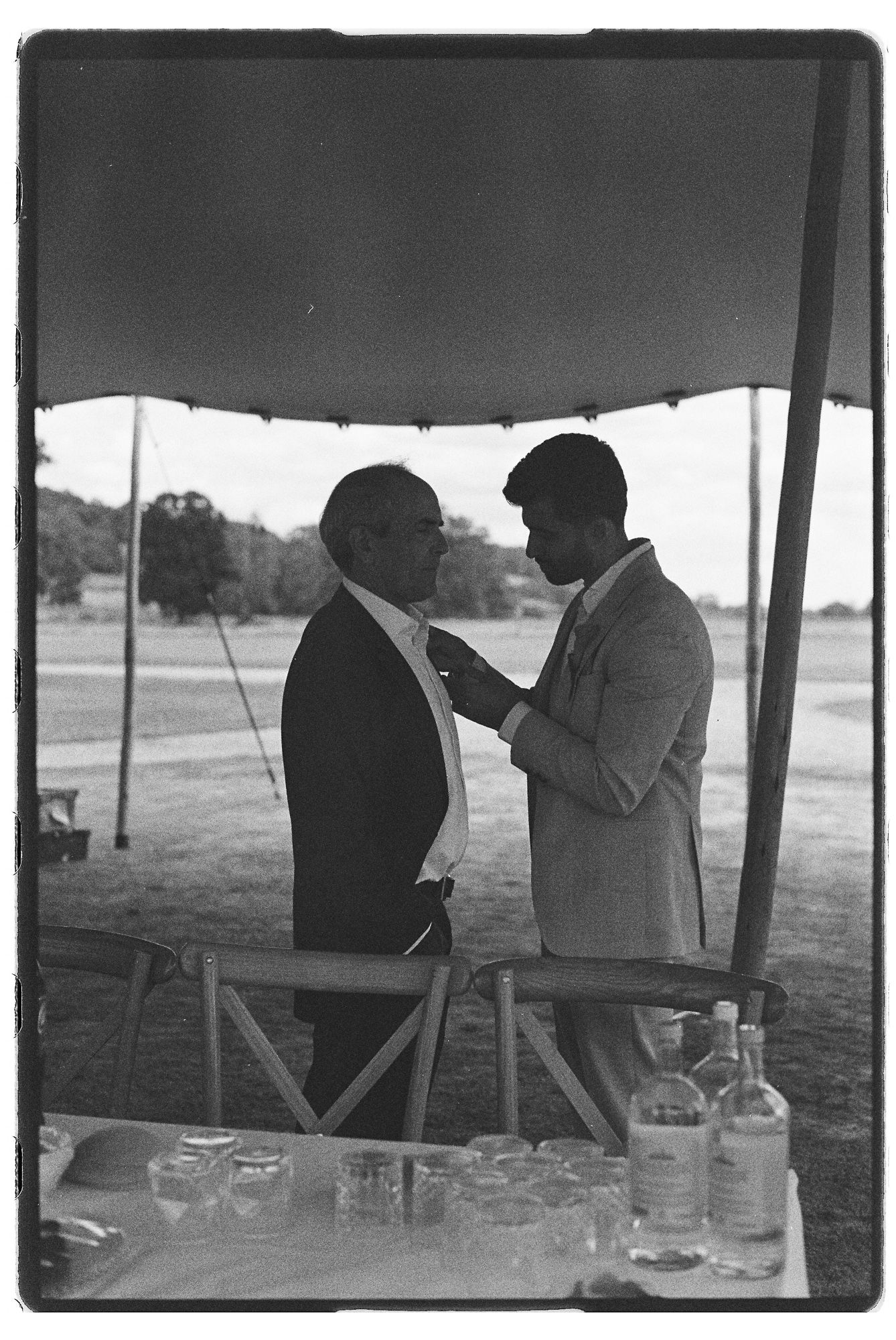 Black and white photo of two men in suits adjusting boutonnieres under a tent at an outdoor wedding reception.