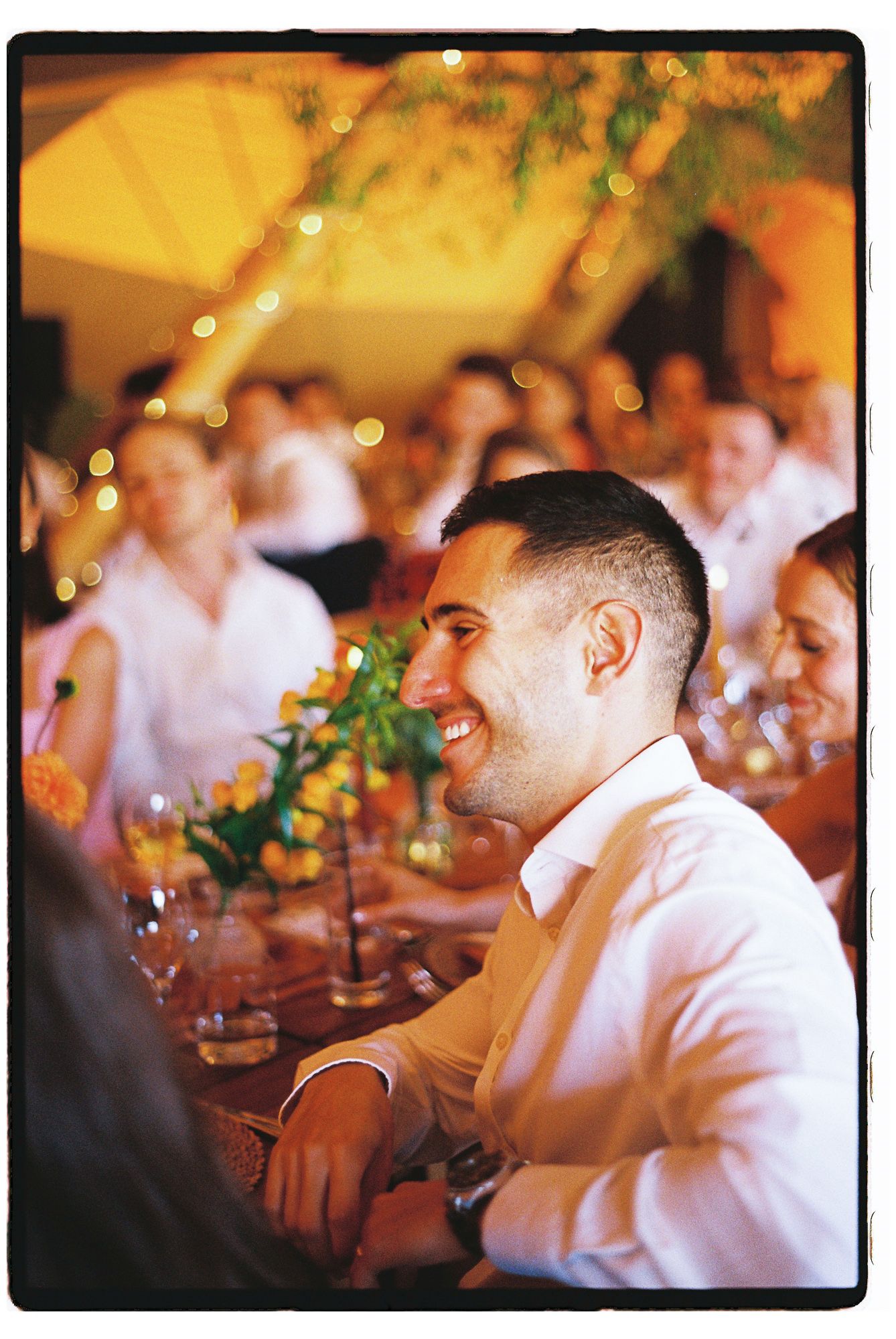 Smiling man in white shirt at festive wedding reception dinner with bokeh lights and floral centerpieces.