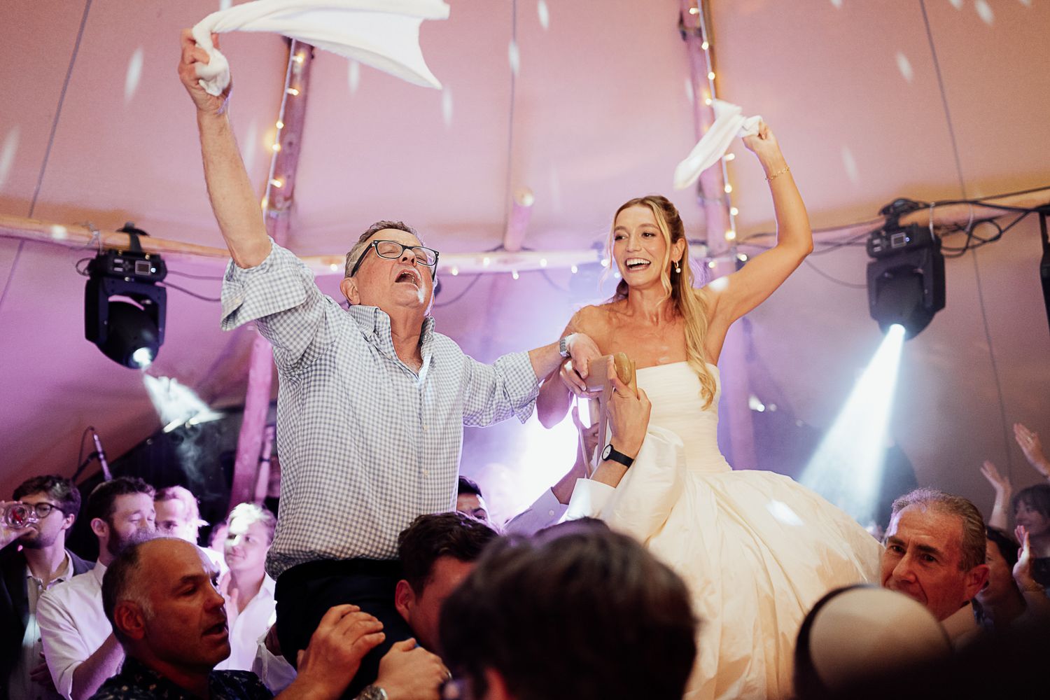 Bride and groom lifted by guests waving napkins during joyful Jewish wedding reception under tent.