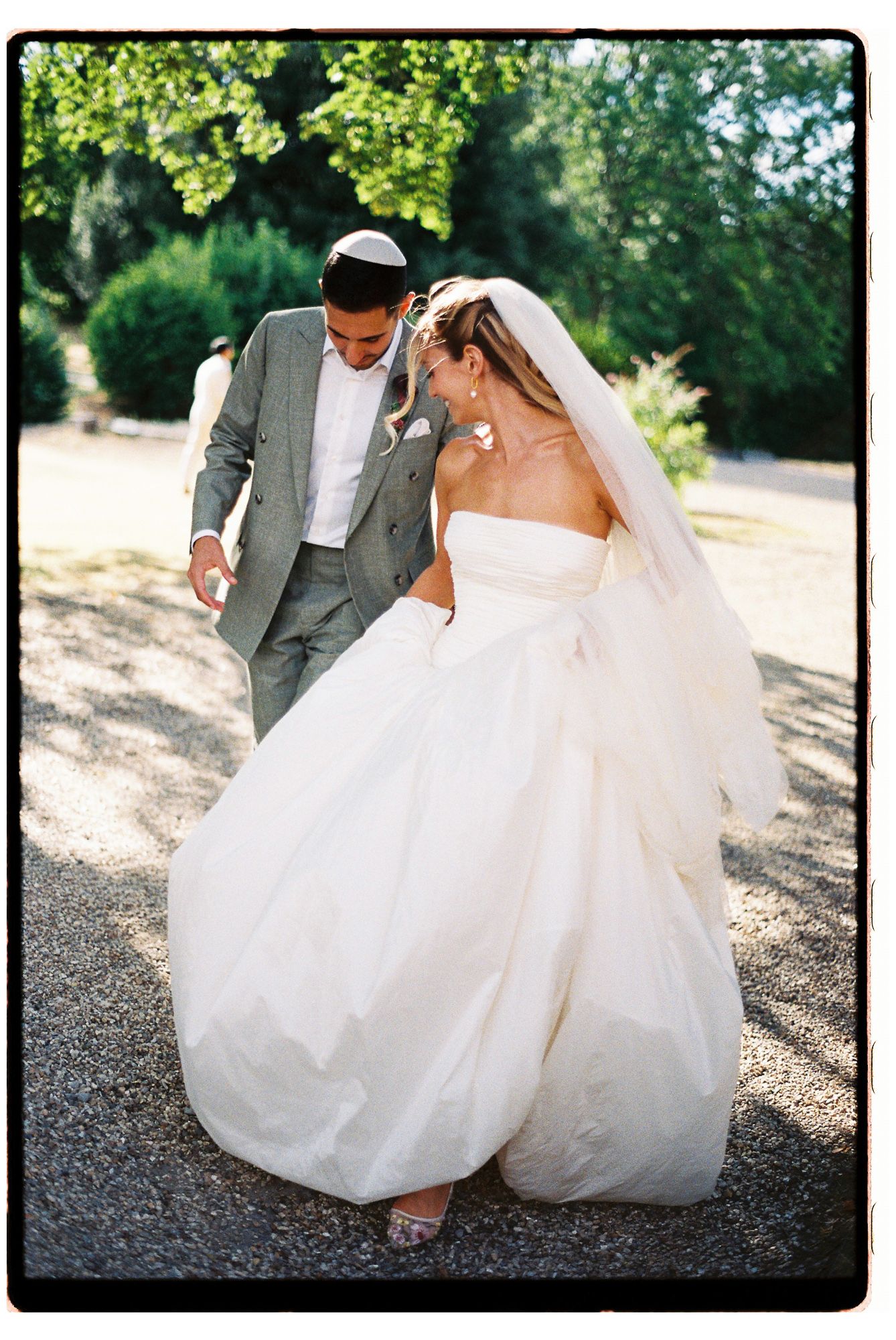 Jewish bride and groom share a romantic moment outdoors, bride in strapless white gown with veil, groom in gray suit with kippah.