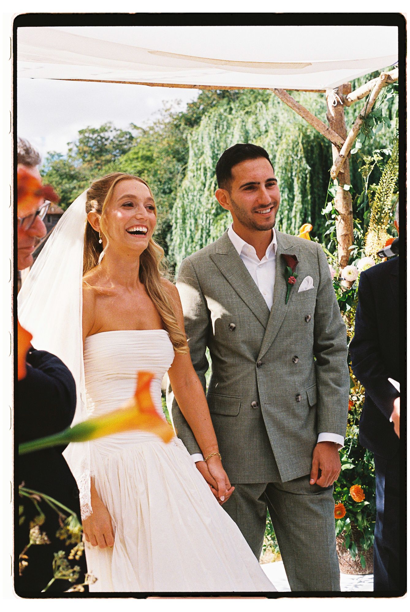 Happy bride and groom holding hands at outdoor wedding ceremony, bride in white strapless gown with veil, groom in gray suit.
